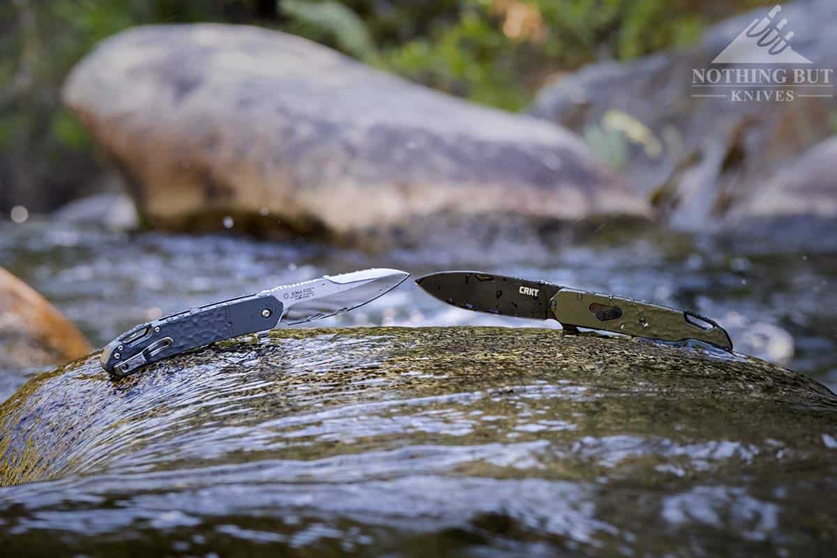 The CRKT Bona Fide Silver and OD Green pocket knives on a rock in the middle of a mountain creek. 