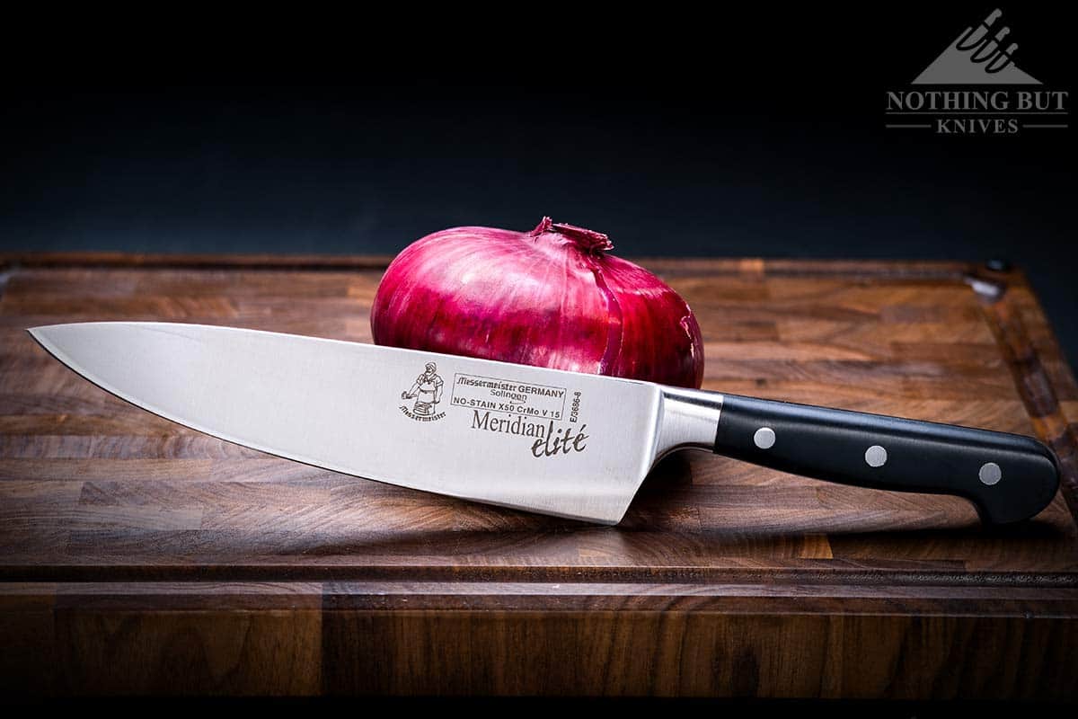 The 8 inch Mesermeister Meridian Elite chef knife next to a red onion on a wood cutting board. 