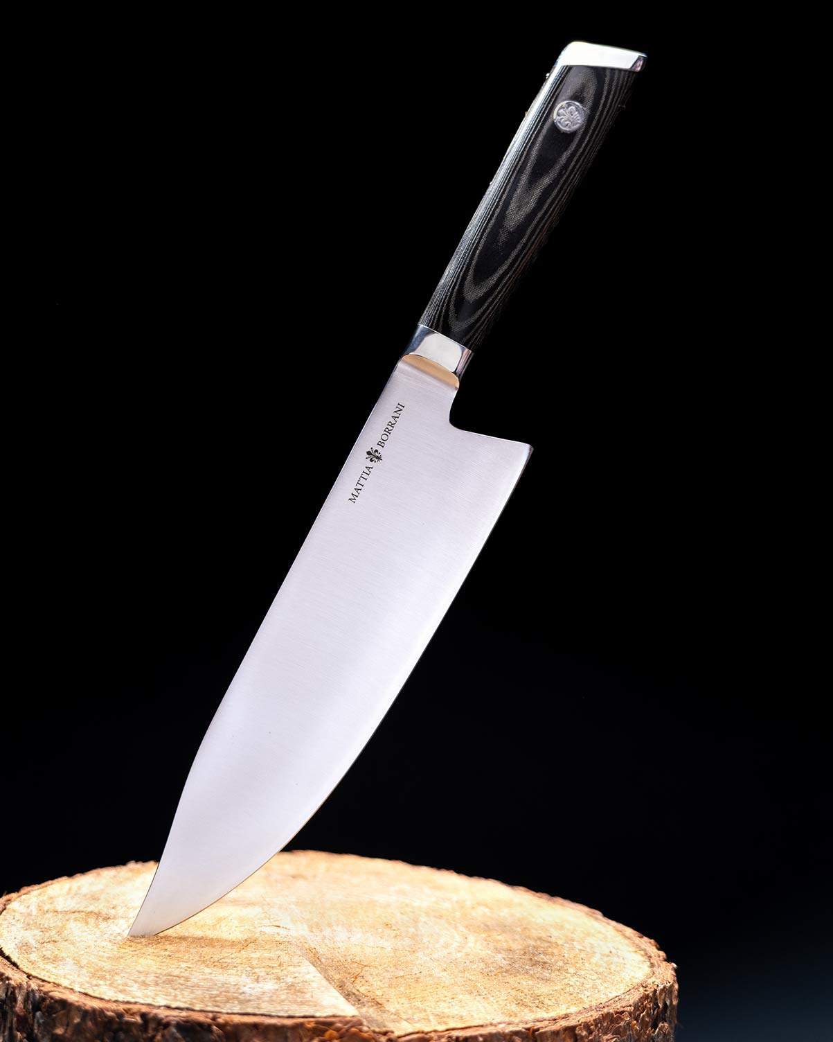 The Mattia Borrani chef knife is a true American style chef knife. It is shown here sticking out of a treestump in front of a dark background.