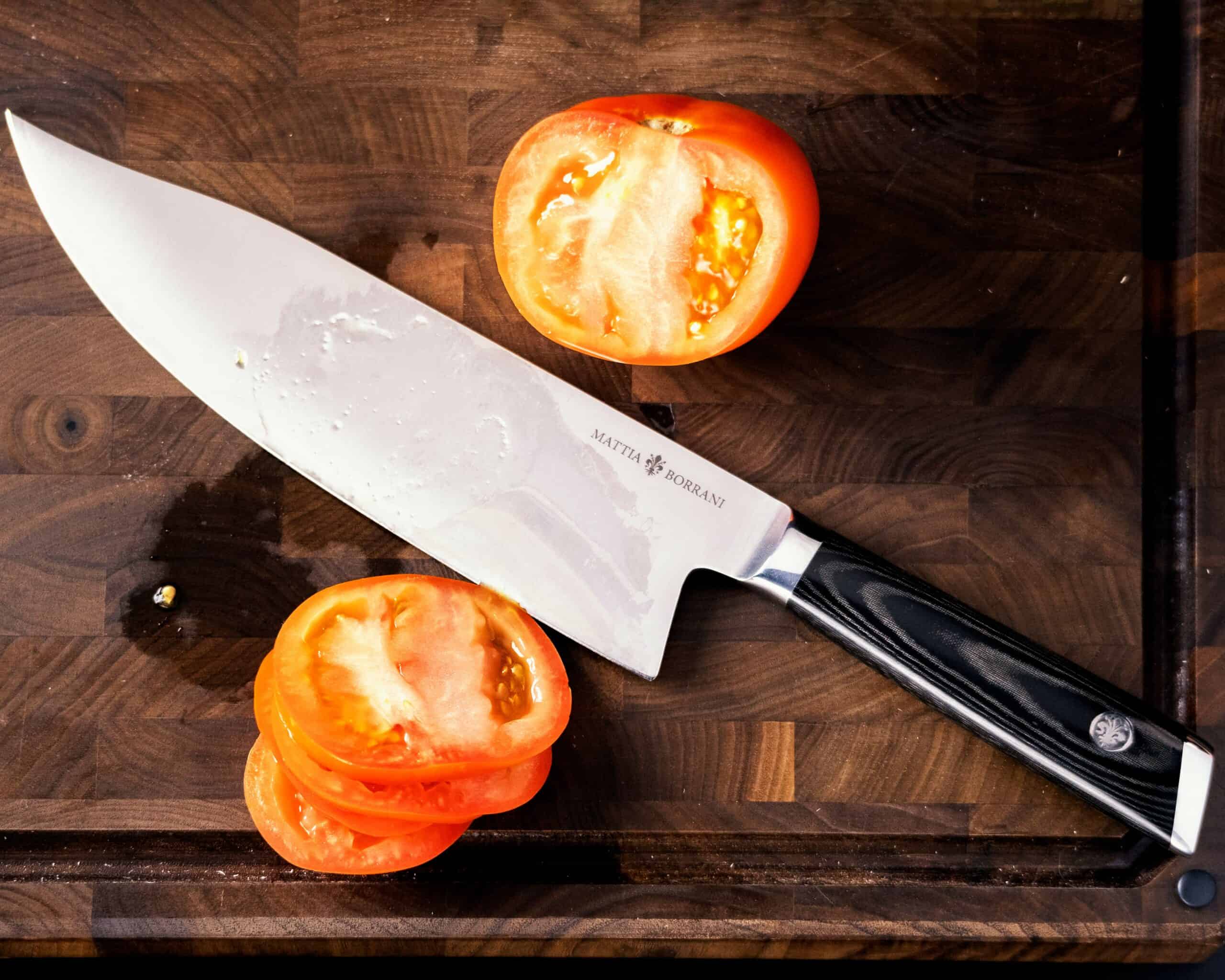 A flat style image showing the Mattia Borrani Bowie chef knife between tomato slices and half a tomato on a dark wood cutting board. 