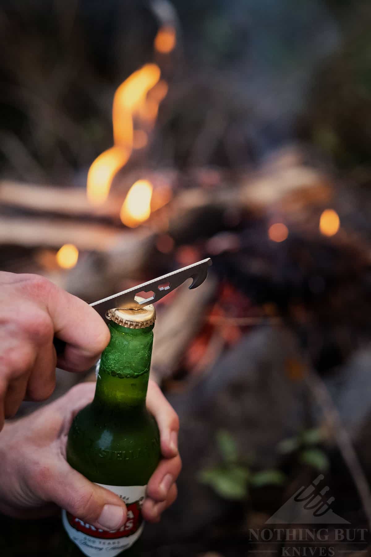 A beer bottle being opened with the Crit's tool bar by a campfire. This illustrates our favorite use of the Civivi Crit in this review. 