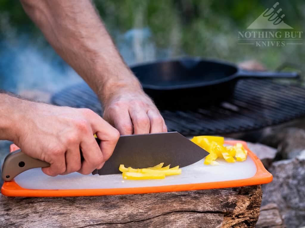 Our Search For The Best Camping Chef Knife - Image 3 The Off-Grid Grizzly V2 chef knife cutting a yellow bell pepper on a plastic cutting board in front of a cast iron pan on a campfire.