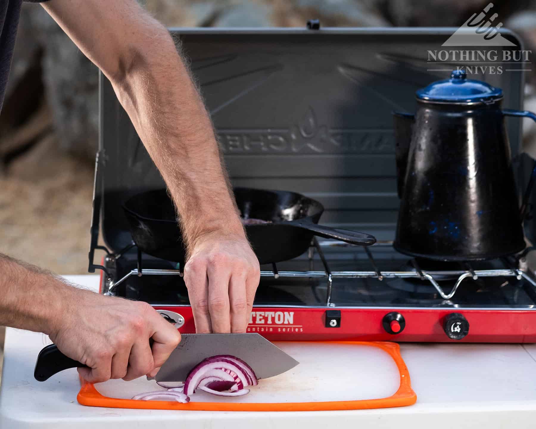 Our Search For The Best Camping Chef Knife - Image 4 Slicing an onion with the Off-Grid Grizzly in front of a camp stove. with a cast iron skillet and a coffee pot.