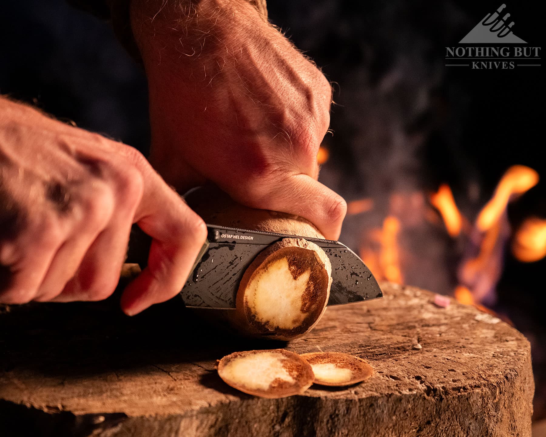 Our Search For The Best Camping Chef Knife - Image 9 A close-up of a potato being sliced with the Civivi Hid folding knife on a stump next to campfire in a forest.