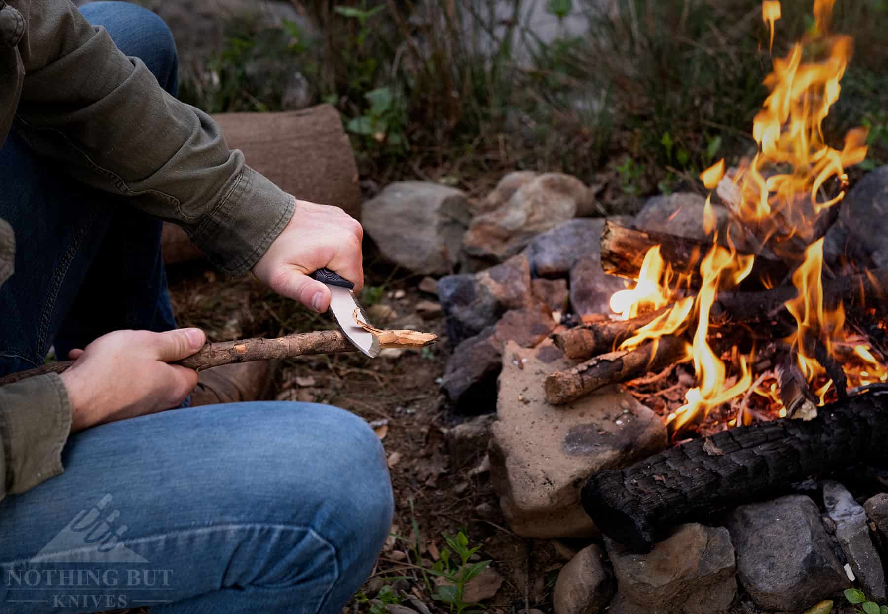 A close up of a person carving a stick next to a campfire to show how effective the scandi grind on the Ridgeback is.