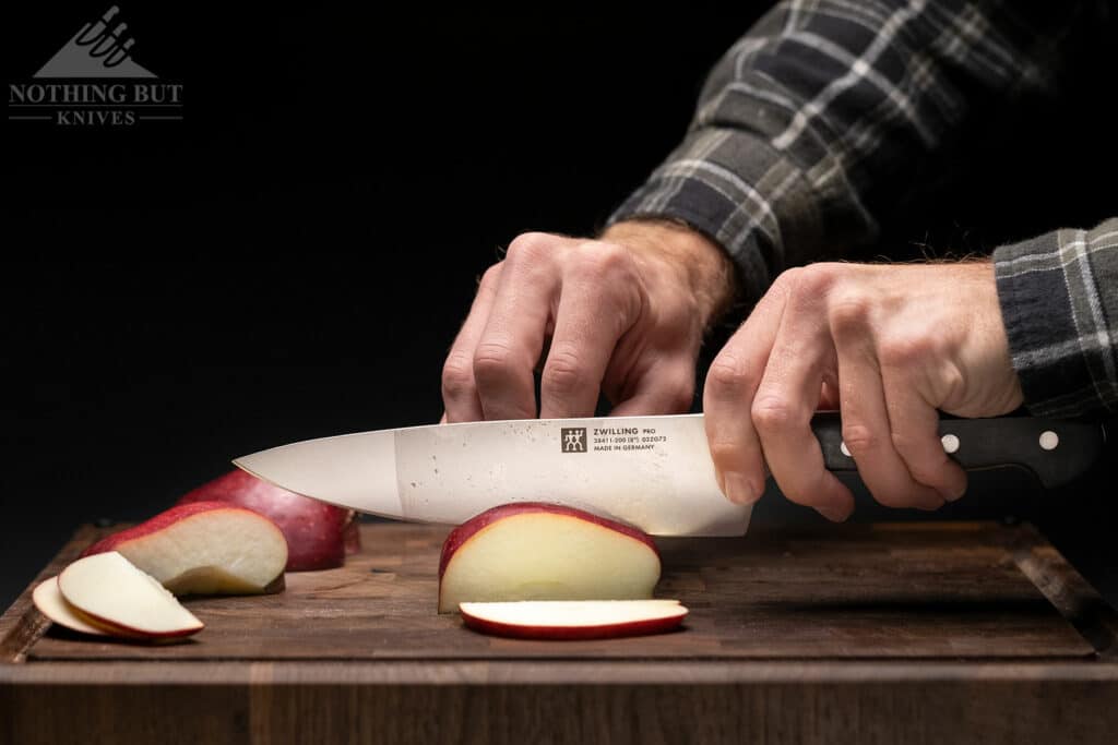 Best High-End Knife Sets For Professional Or Home Use - Image 10 The Zwilling 8-inch Pro chef knife being used to slice a red apple on a dark wood cutting board.