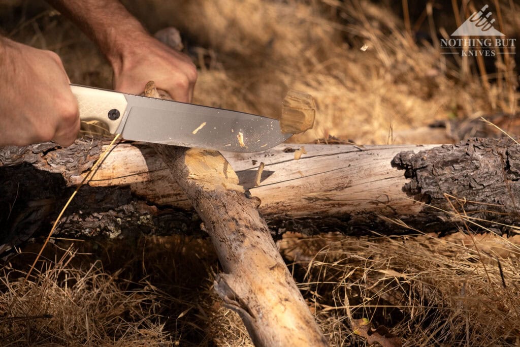 Top Survival Knife Beatdown - Hands On Testing - Image 10 A close-up of the Off-Grid Tracker XXL being used to chop a branch from an oak tree in a field of brown grass.