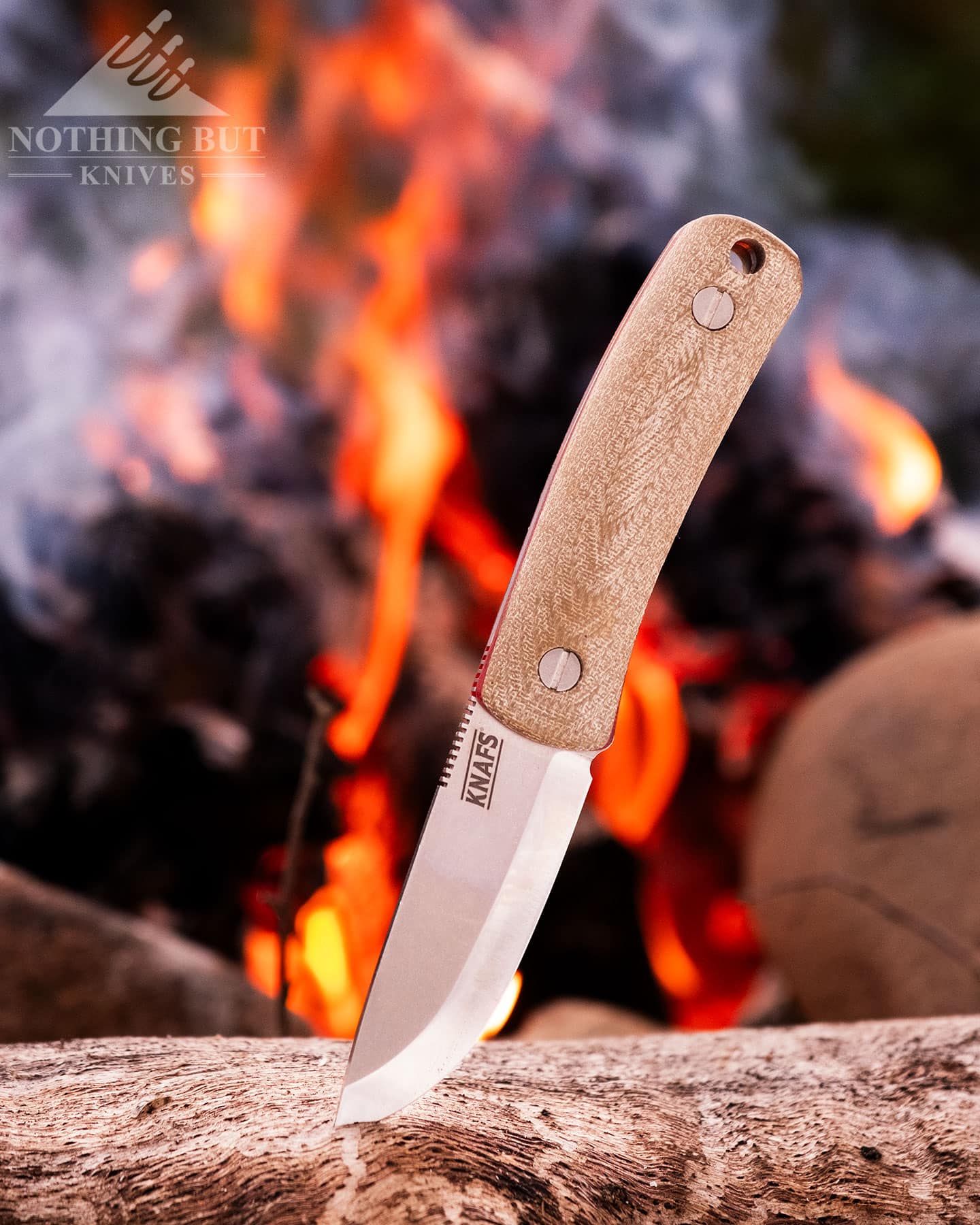 A close-up of the Knafs Lulu Bushcraft knife sticking out of a log next to a campfire in a forest. 