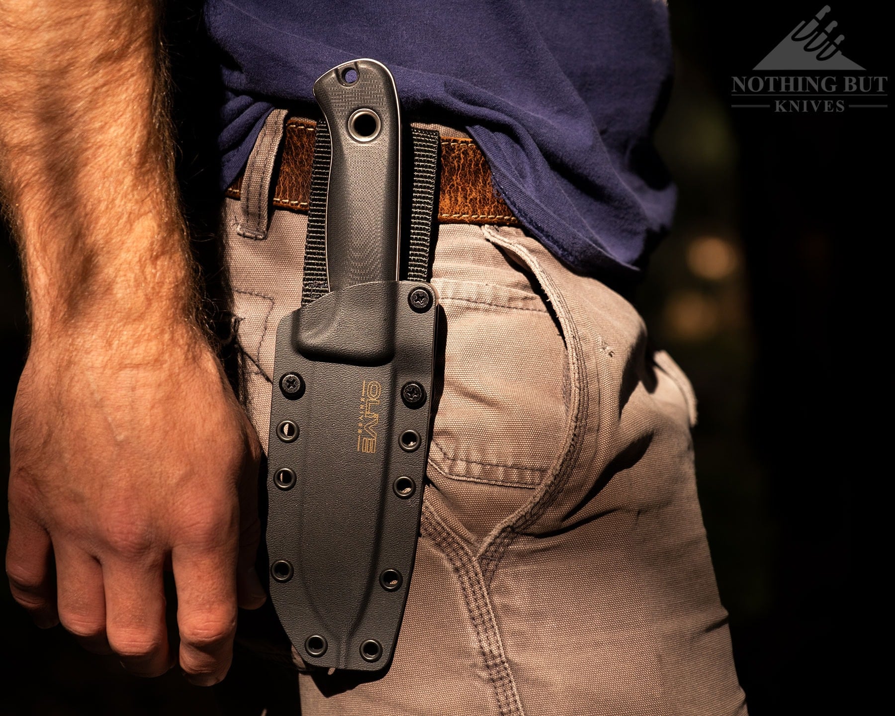 The Olive Arbor bushcraft knife in its pancake style kydex sheath attached to a person's belt.