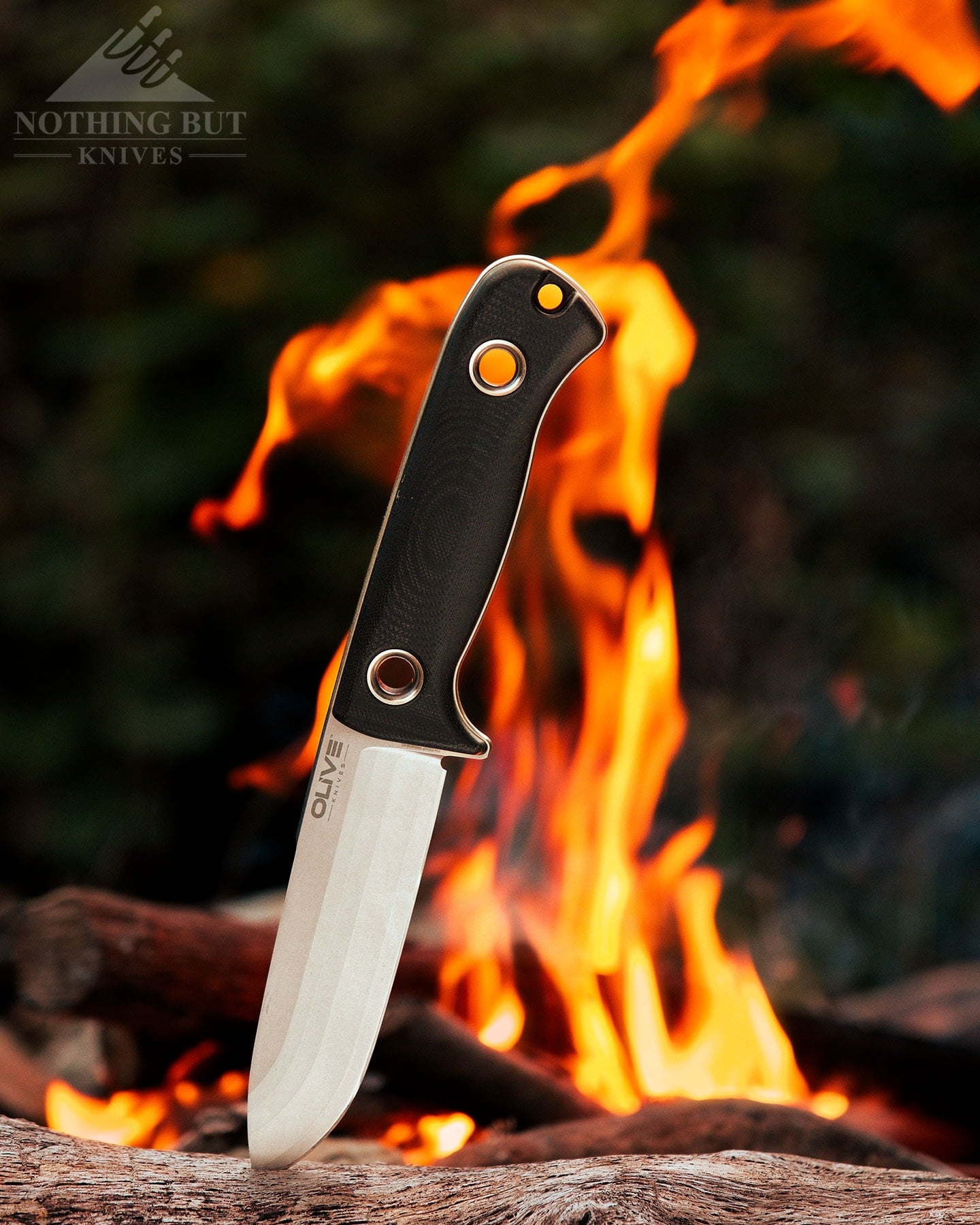 The Olive Arbor fixed blade shown here with its blade tip sticking out of a log next to a campfire in the woods. 