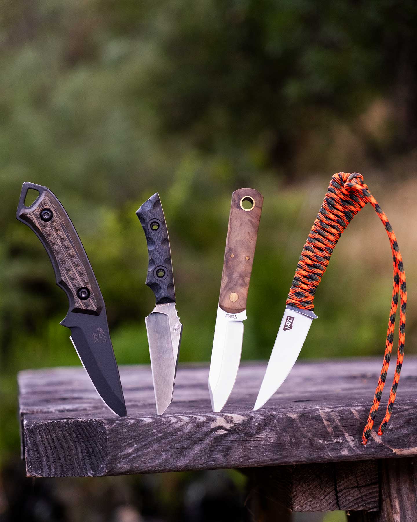Four of the top EDC fixed blade knives with their blades sticking out of a picnic table in front of a green forest background outdoors.