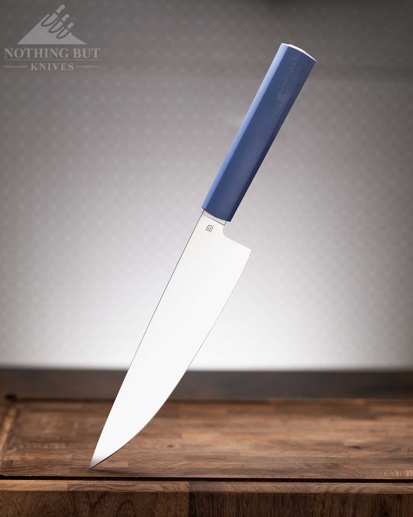 Review Of The Butterfork 4 Piece Kitchen Knife Set - Image 1 A blue handled Butterfork chef knife shown here handle up and blade down on a dark wood cutting board in front of an off-white background.