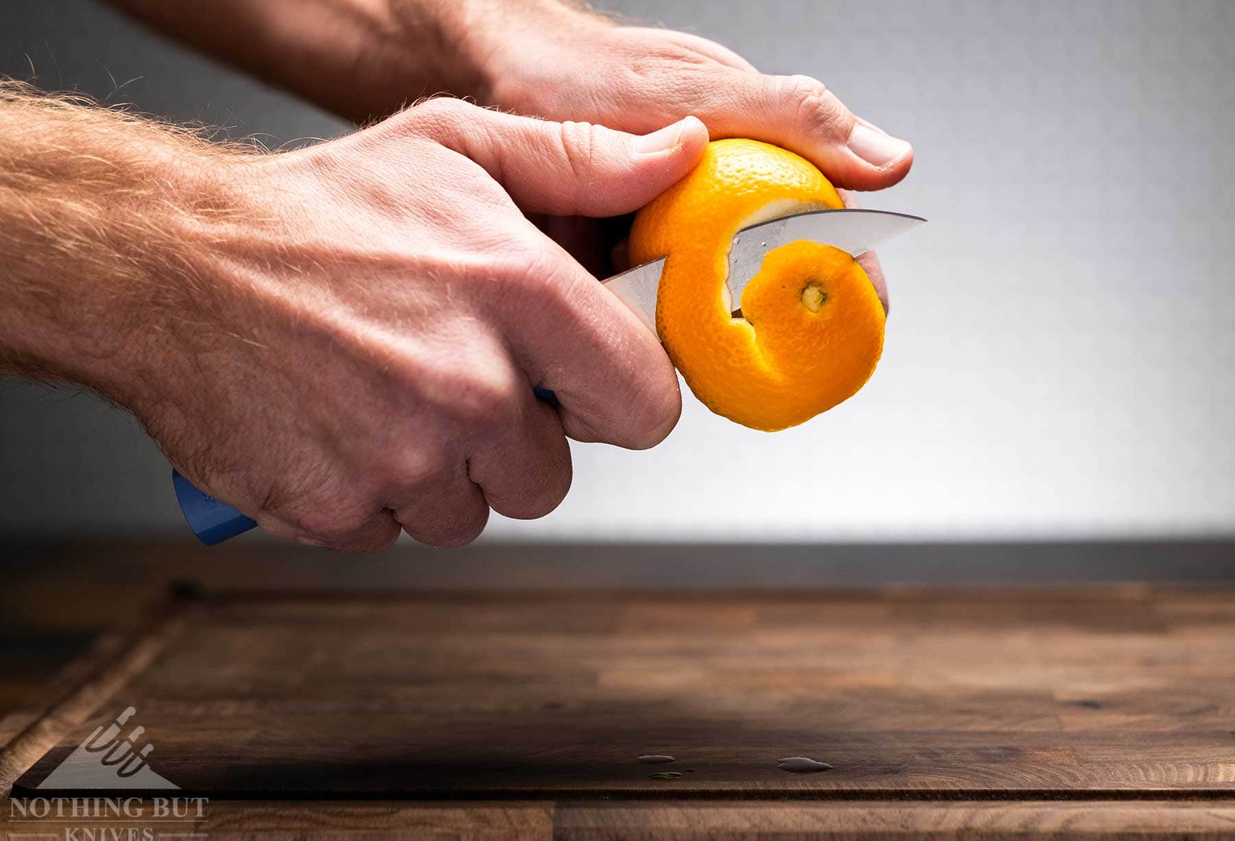 Review Of The Butterfork 4 Piece Kitchen Knife Set - Image 6 The Butterfork paring knife halfway through sprial peeling an orange above a wood cutting board in front of an off-white background.