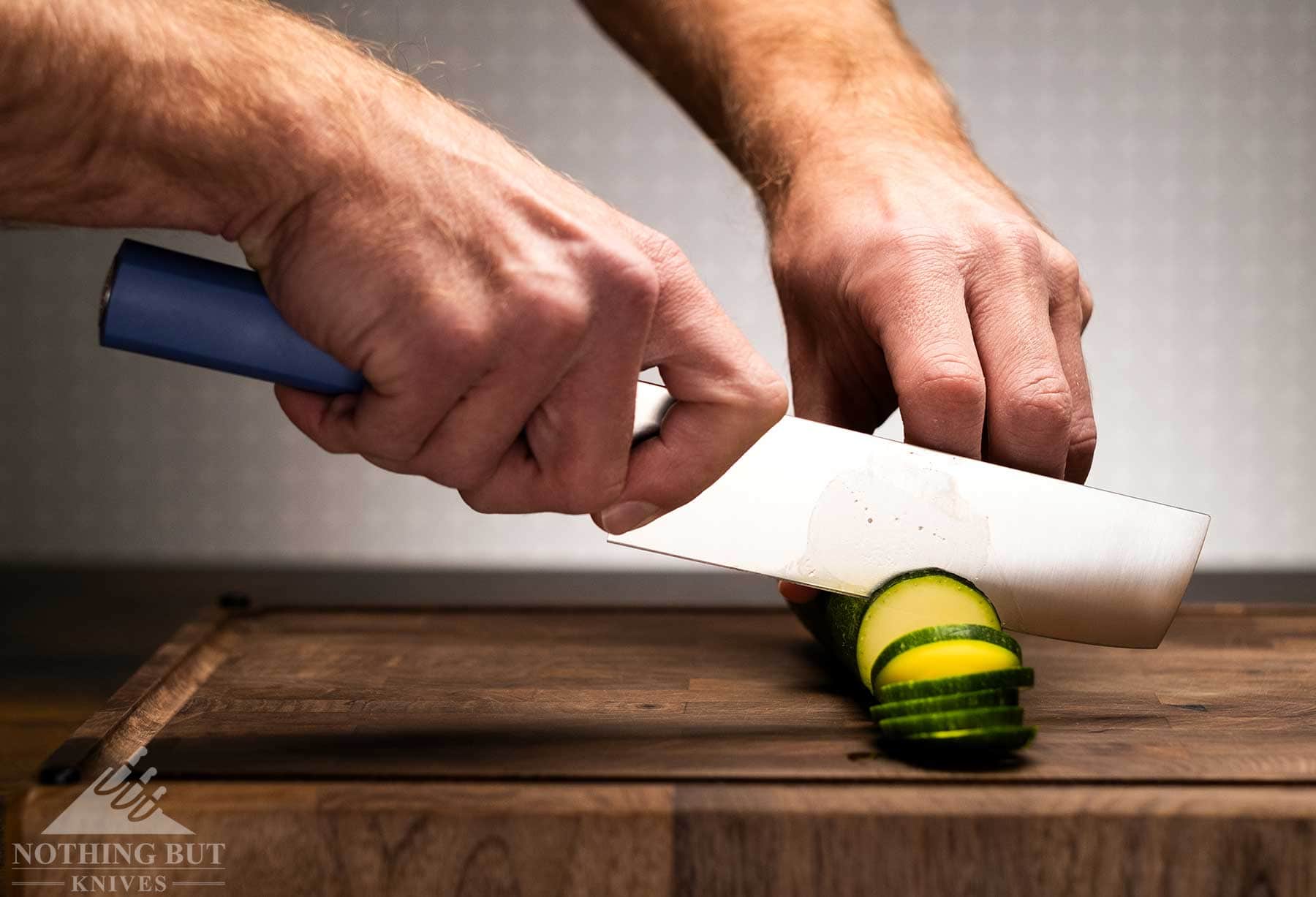 Review Of The Butterfork 4 Piece Kitchen Knife Set - Image 4 A Butterfork Nakiri knife with a blue handle being used to cut a cucumber into slices.