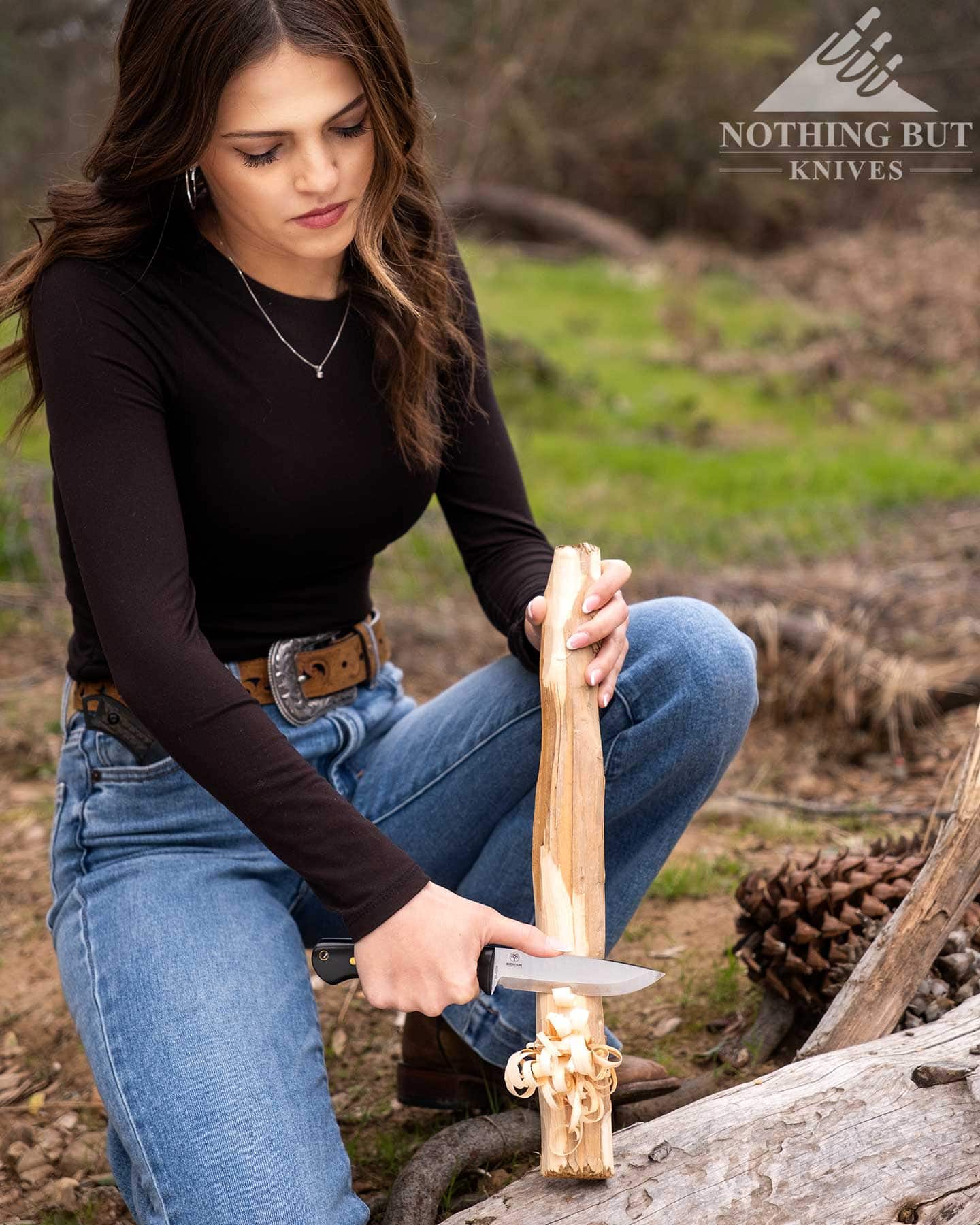 Boker Dbk Bushfriend 2000 Review - Image 7 A young woman in jeans and a black long sleeve shirt carving a small log in the forest.