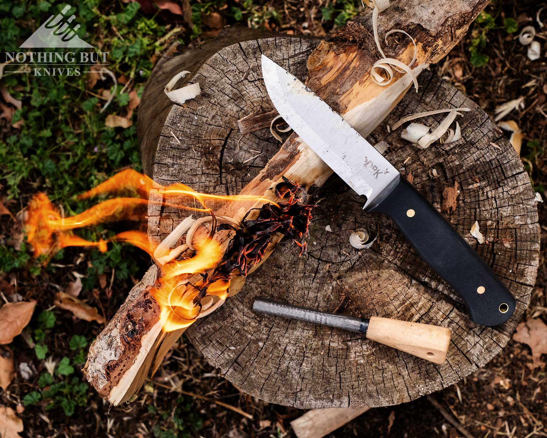 Boker Dbk Bushfriend 2000 Review - Image 14 An overhead view of a bushcraft knife and ferro rod next to a burning stick on top of a piece of firewood.