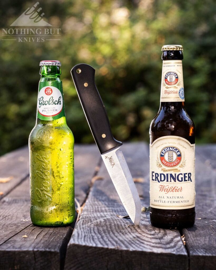 Boker Dbk Bushfriend 2000 Review - Image 2 The Boker DBK Bushfriend 2000 Scandi sticking out of a picnic table between a bottle of German beer and a bottle of Dutch bee.