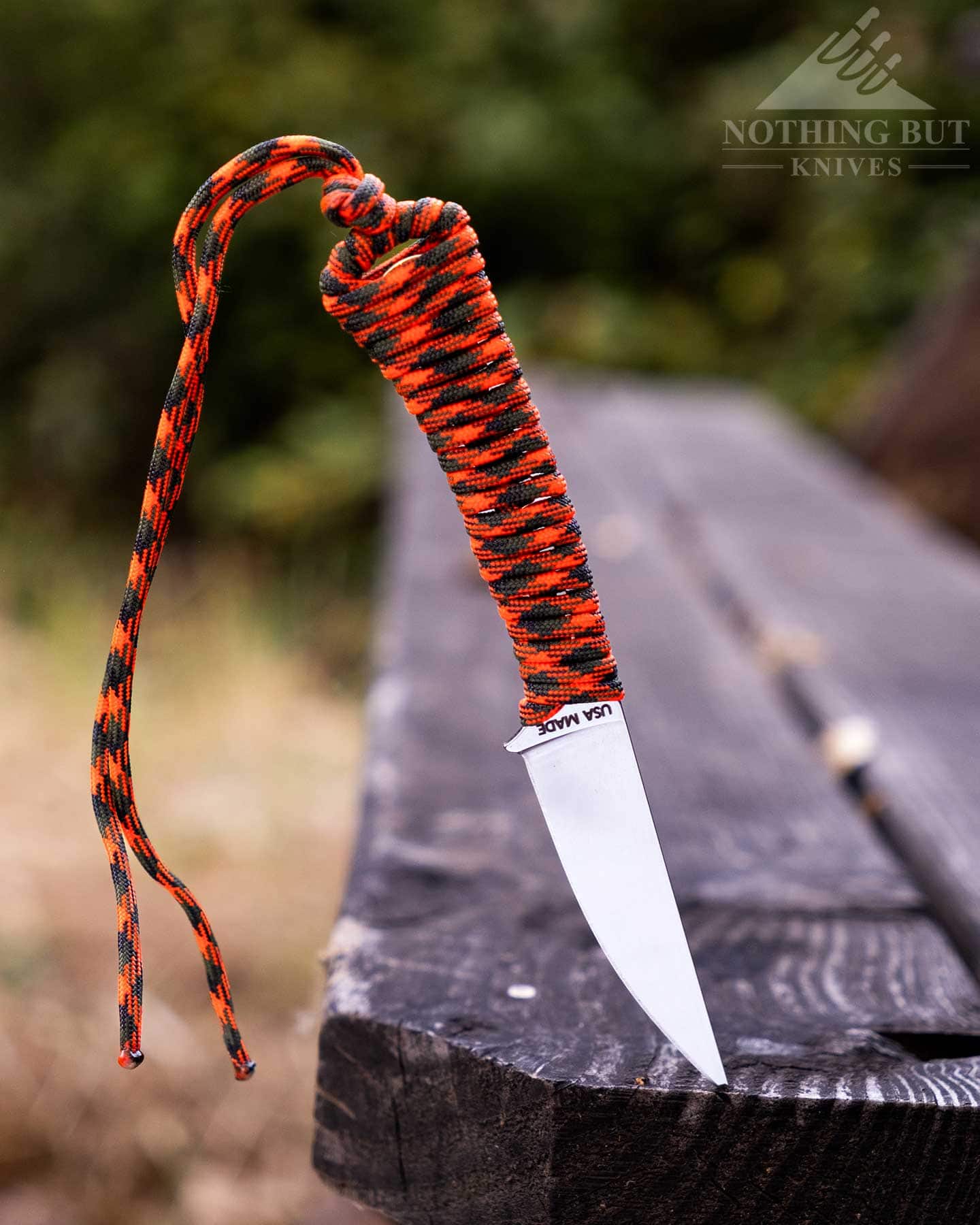 Our Favorite Fixed Blade Edc Knives And How To Carry Them - Image 12 The MKC Speedgoat 2.0 with an orange paracord covered handle sticking out of a picnic table bench in front of a blurry green background.