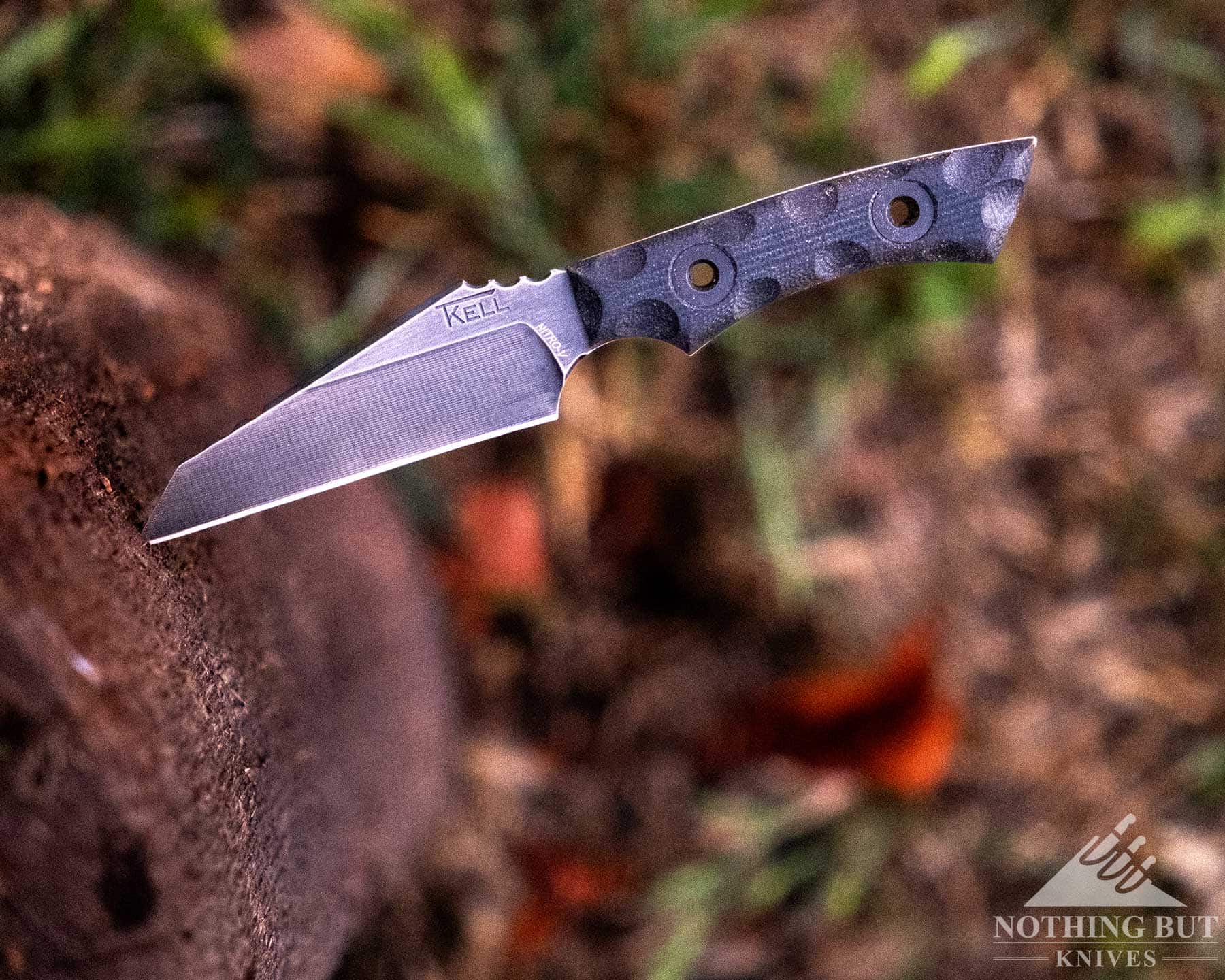T.kell Wharnedc Review - Image 15 An overhead view of a small knife with its blade sticking out of a piece of firewood above a grass-covered field.