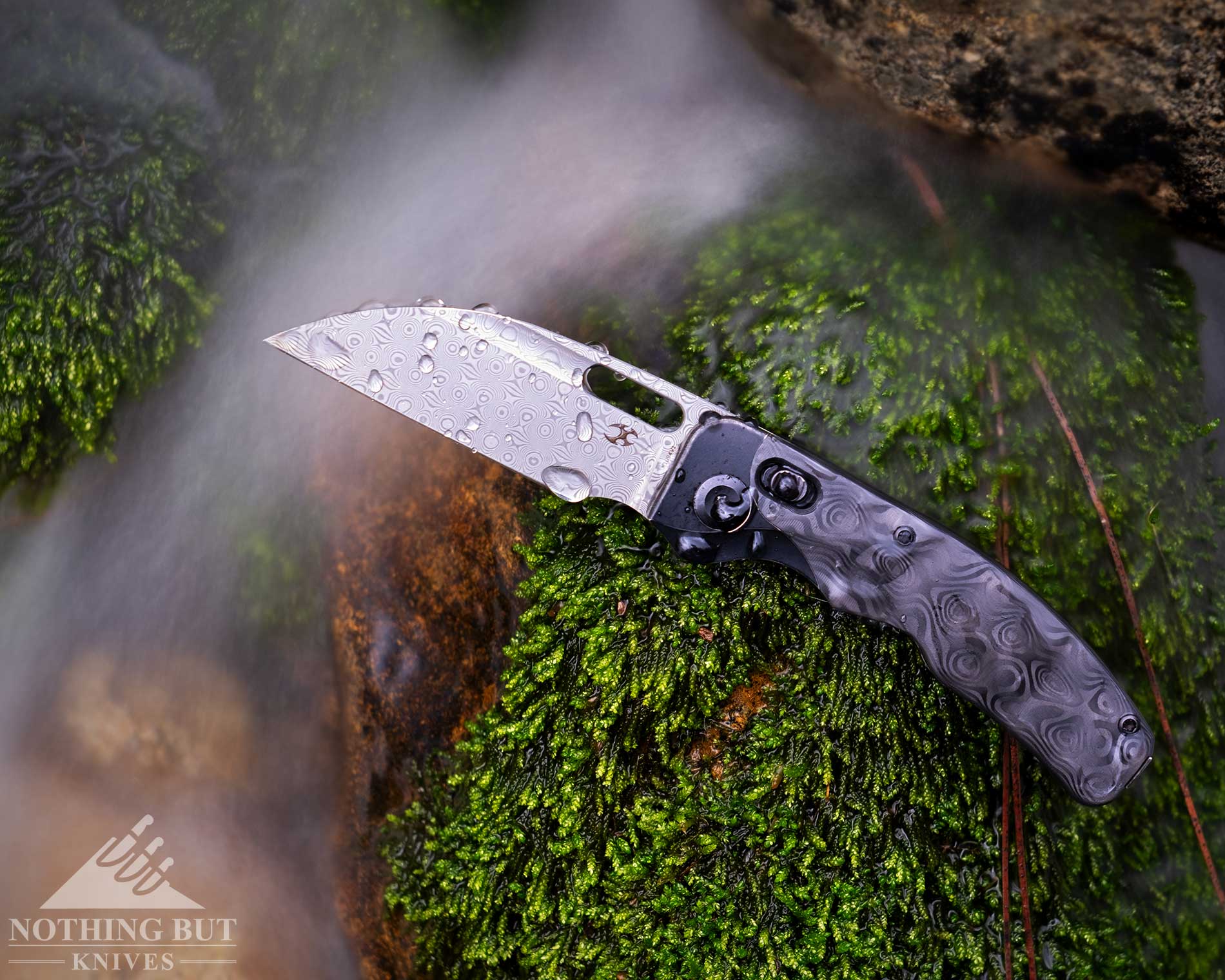Kansept Vivid Review - Image 9 An overhead view of the Kansept Vivid pocket knife with its Damascus steel blade in the open position on top of a mossy rock next to a creek.