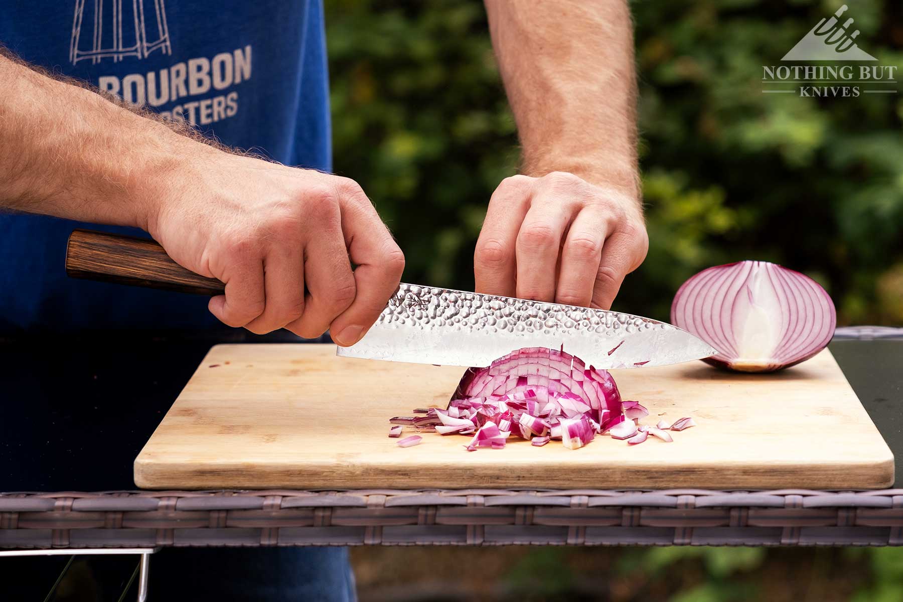 Demko Gyuto Chef Knife Review - Image 6 The Demko Gyuto chef knife being used to slice a red onion on a Bamboo cutting board outdoors in front of a blurry green background.