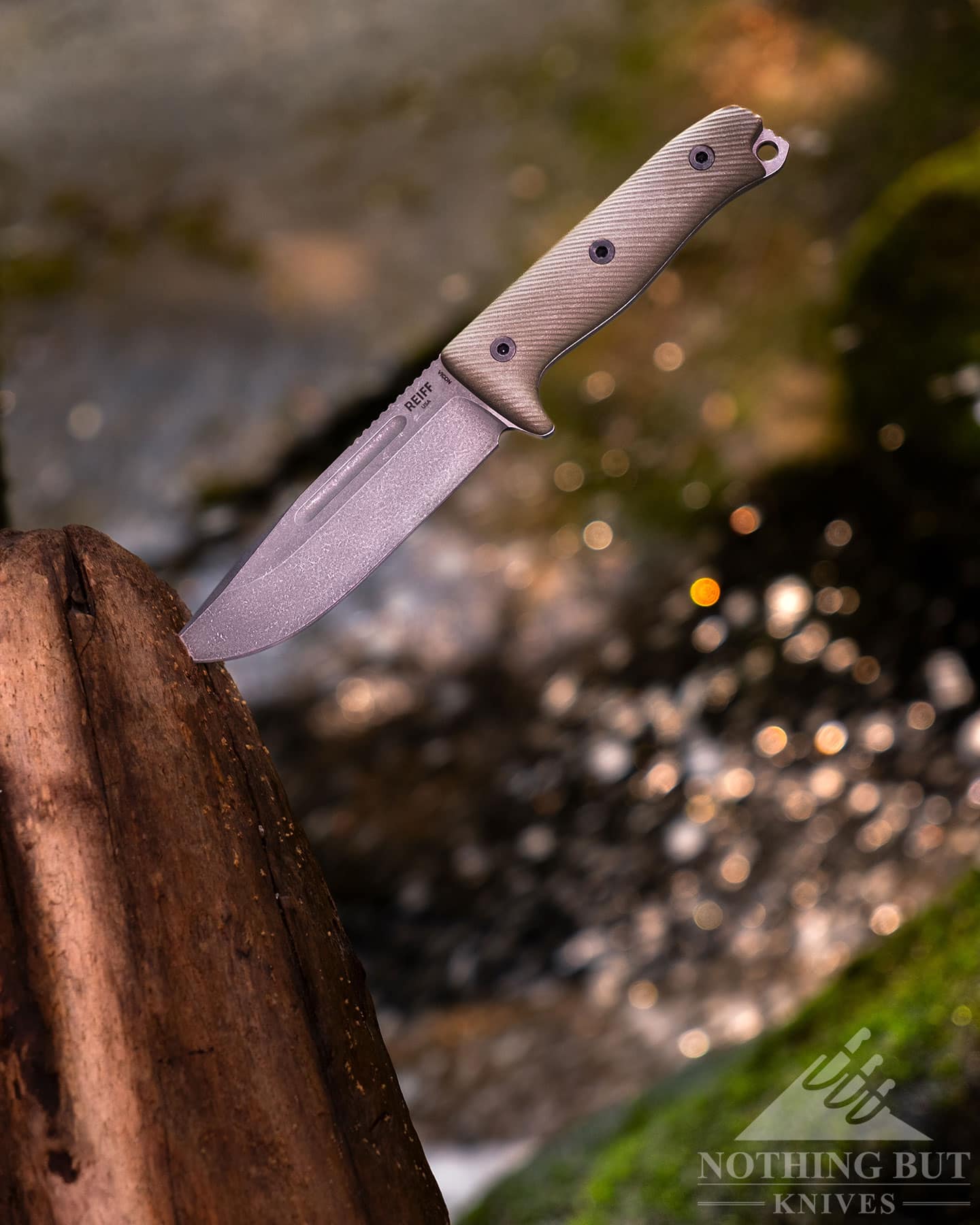 Water splashing behind the Reiff Vicon survival knife which is shown here with its blade tip sticking out of a log. 