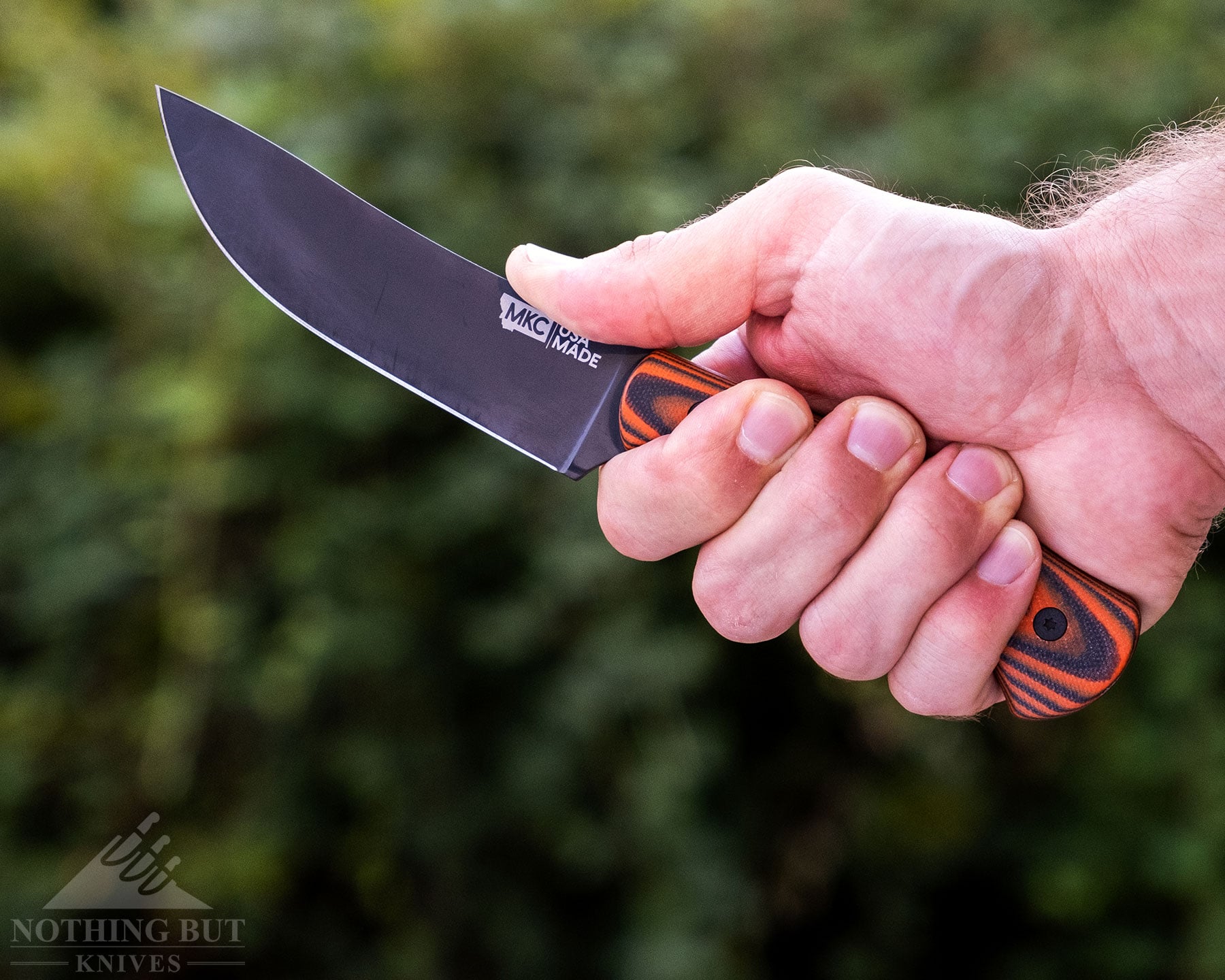 A side view of the Montanan Knife COmpany Stonewall Skinner in a person's right hand in front of a blurry green background. 