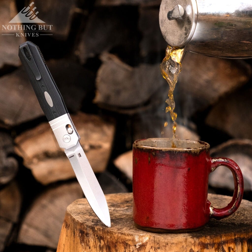 A close--up of the CRKT Redemption pocket knife sticking out of a log next to a coffee cup tha is being filled in front of a wood pile. 