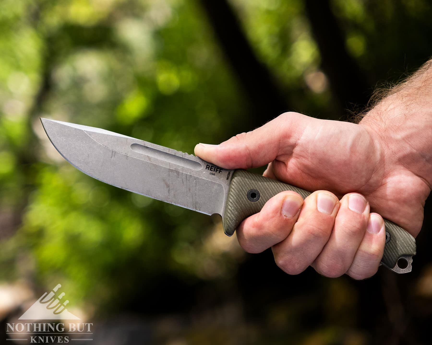 A close-up of the Reiff Vicon being held in a person's right hand in front of a forest. 