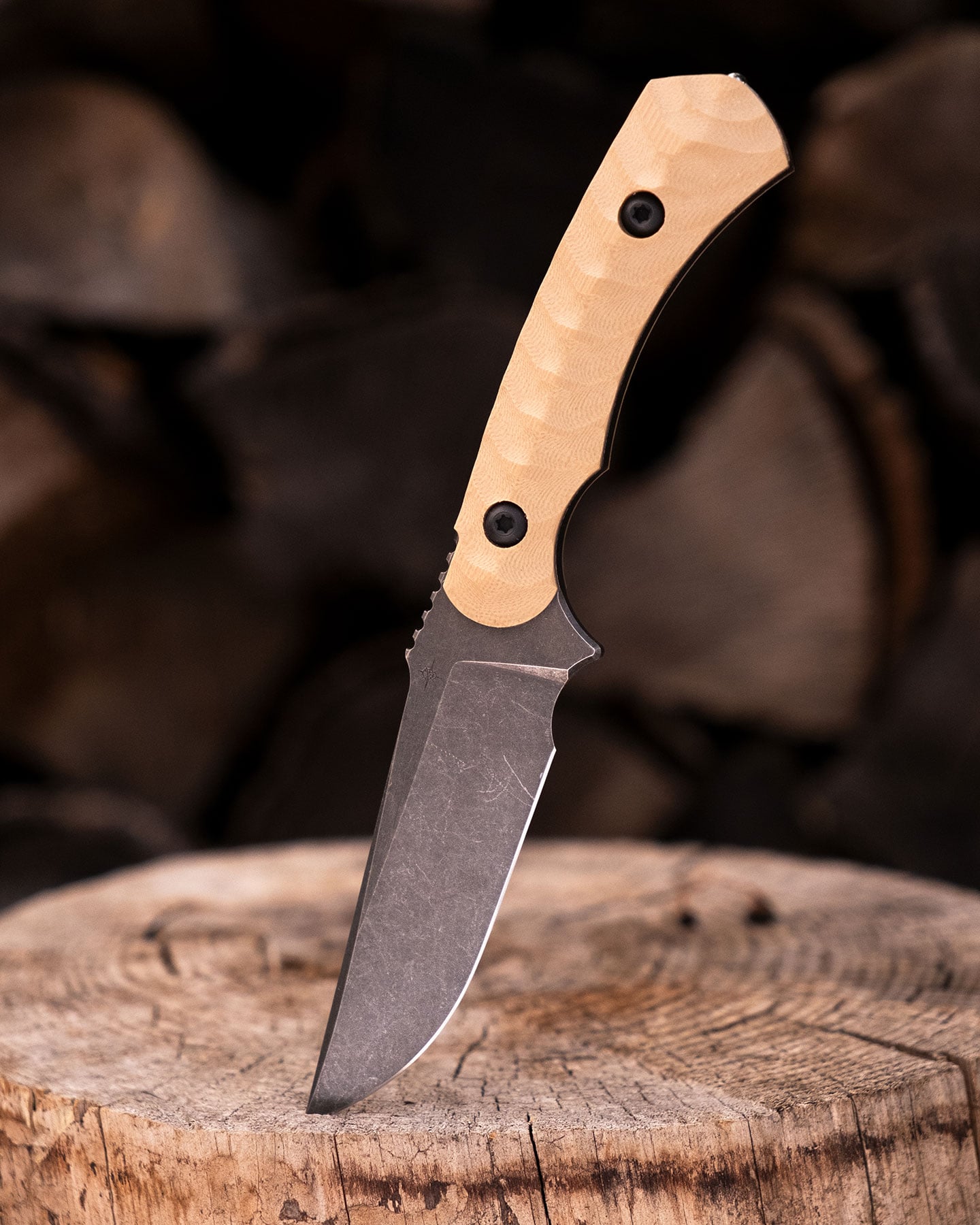 The Toor Brauler fixed blade tactical knife with its blade tip sticking out of a log in front of a stack of firewood.
