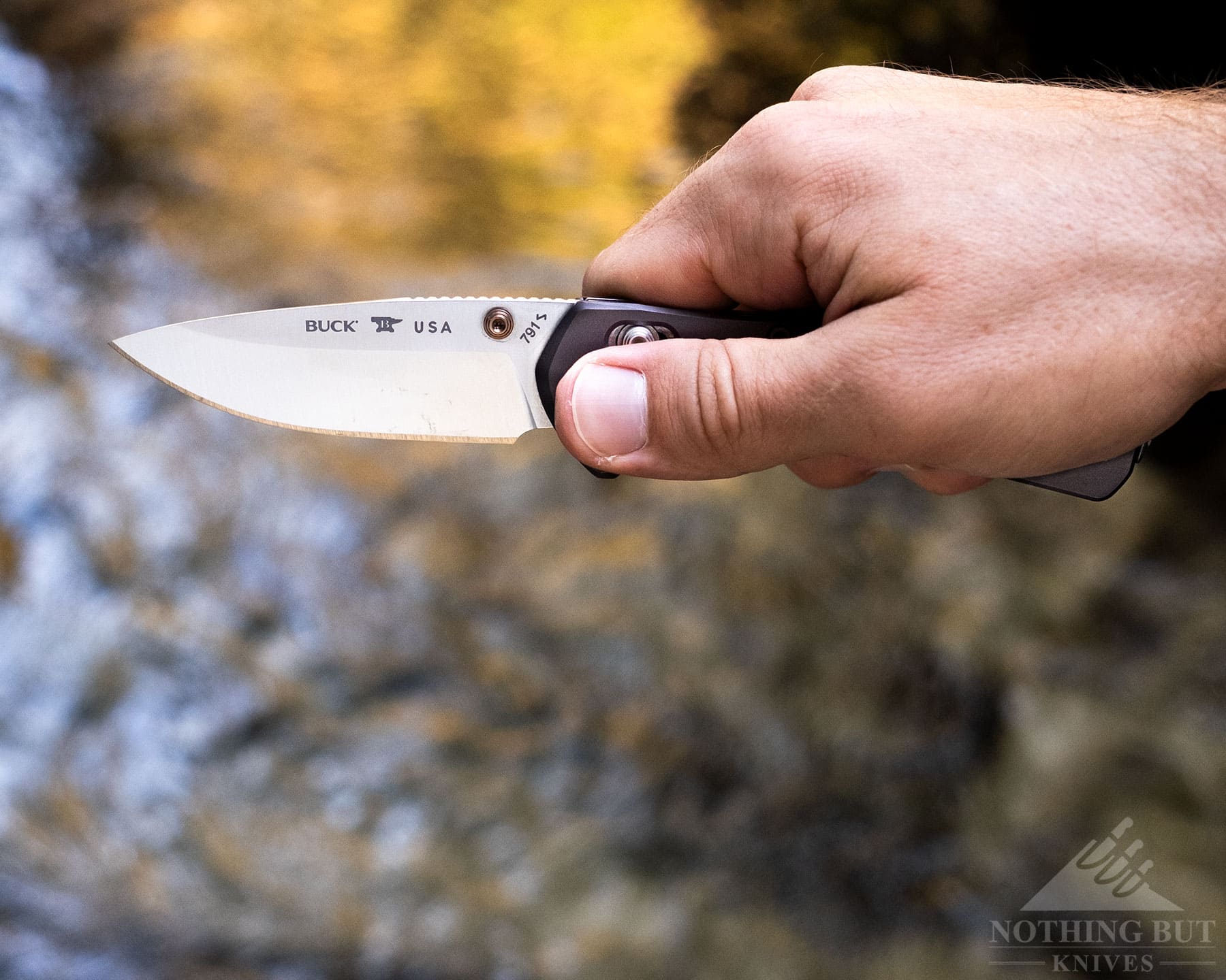 A close-up of a person's thumb actuating a crossbar lock on the Buck Range Elite knife in front of a mountain creek.