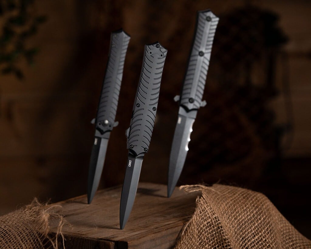 Three different variations of the CRKT Xolotl OTF sticking out of a block of wood that is partially covered in burlap.