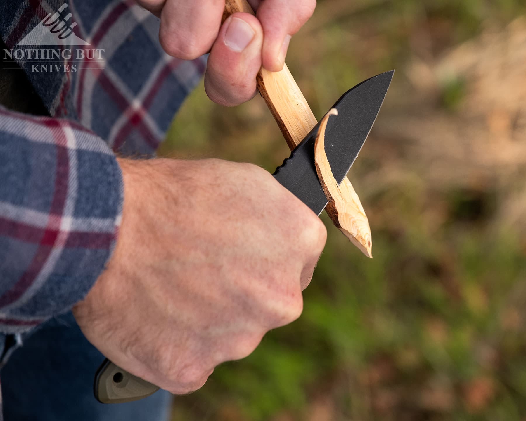 Schwarz Knives Overland X Review - Image 4 A close-up of the Schwarz Overland X being used to carve a stick in front of a blurry forest background.