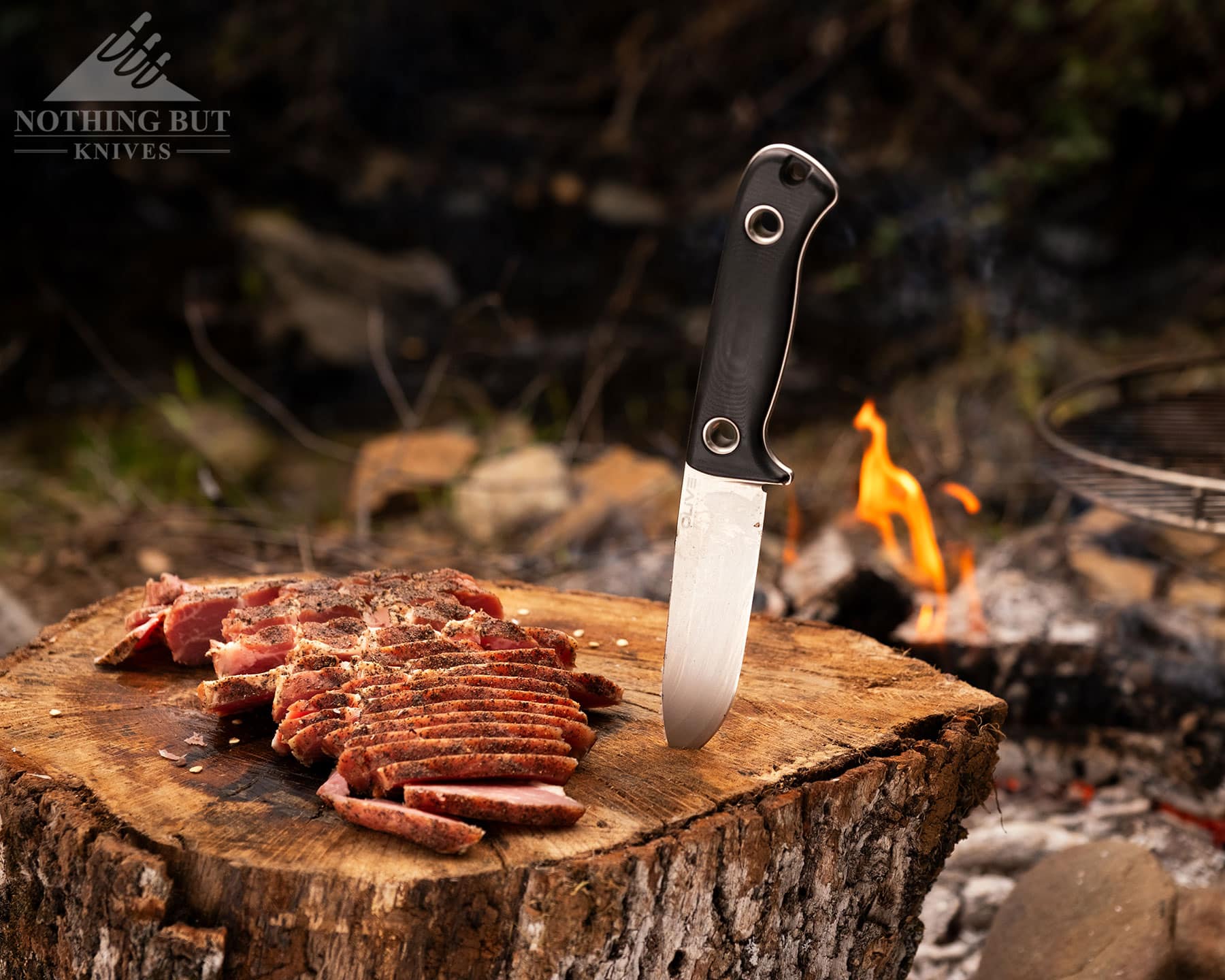 Olive Arbor Review - Image 9 A close-up of the Olive Arbor bushcraft knife next to a slab of sliced Buckboard Bacon in front of a campfire