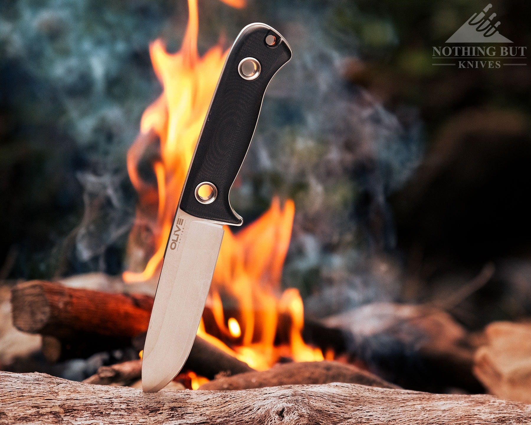 Olive Arbor Review - Image 2 The Olive Arbor knife on a log in front of a campfire in the woods.