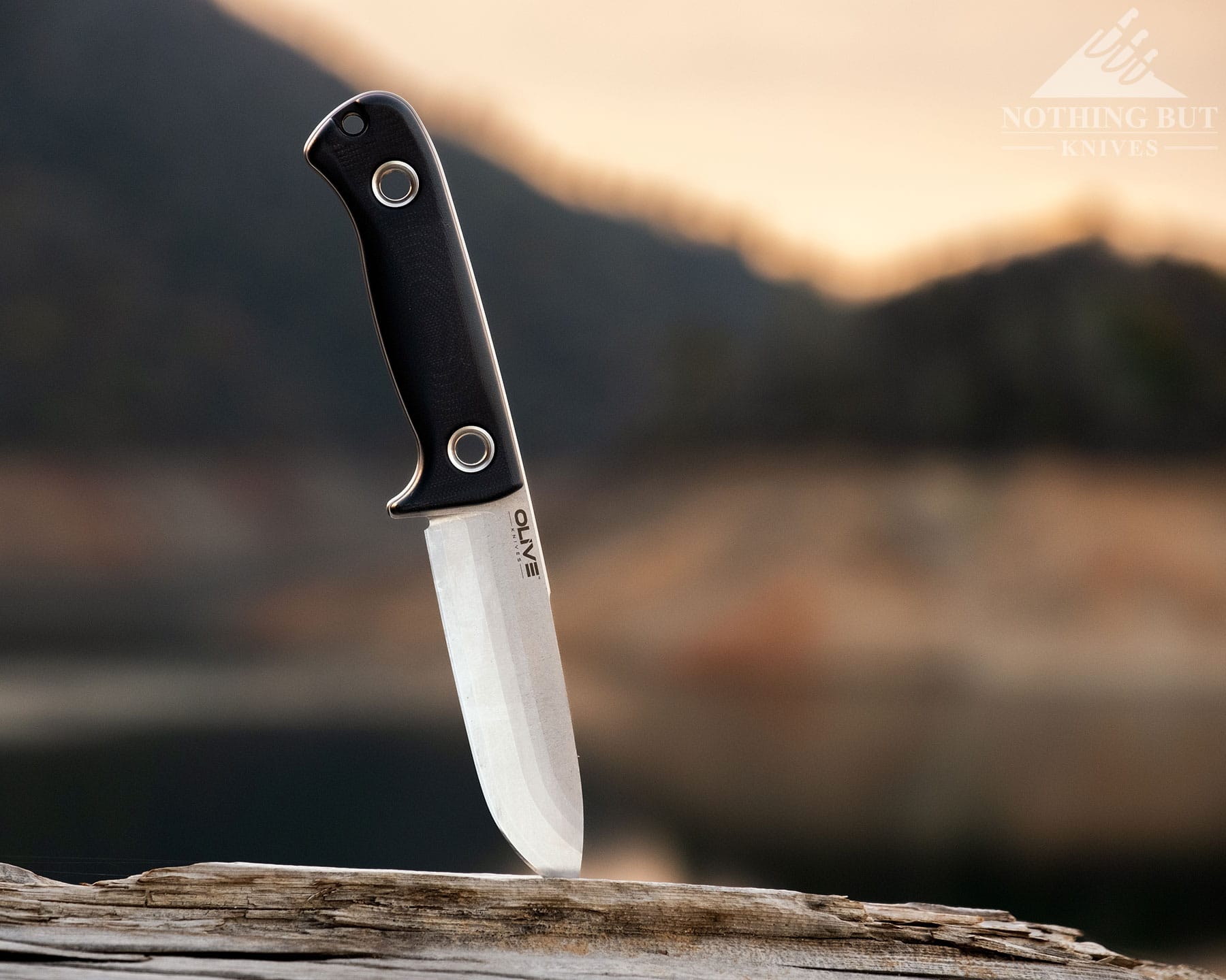 Olive Arbor Review - Image 7 A landscape showing the Olive Arbor bushcraft knife sticking out of a piece of driftwood in front of a mountain lake at sunset.