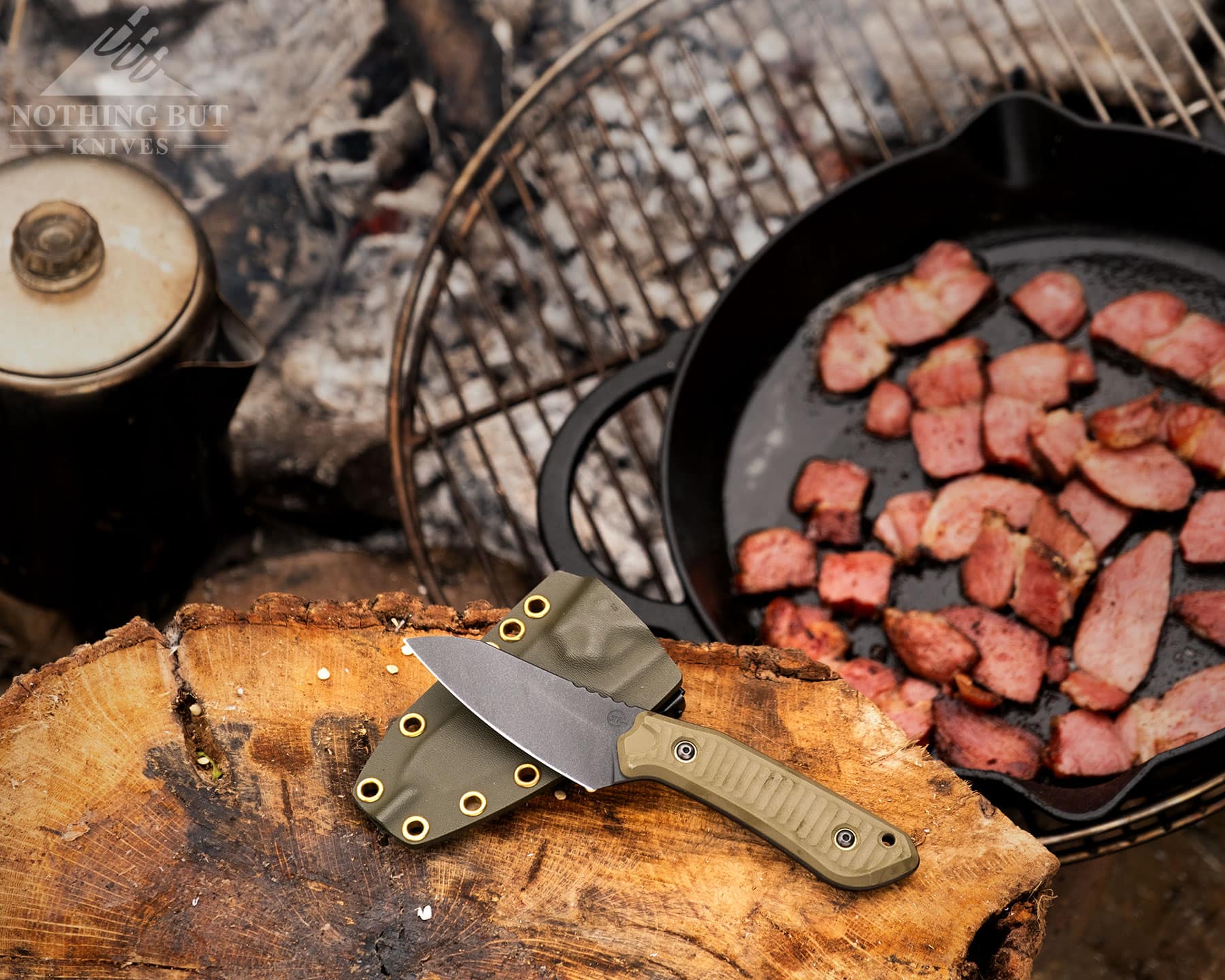 Schwarz Knives Overland X Review - Image 11 An elevated view of the Schwarz Knives Overland X with its kydex sheath on a log next to a campfire with a frying pan full of bacon next to a percolator.