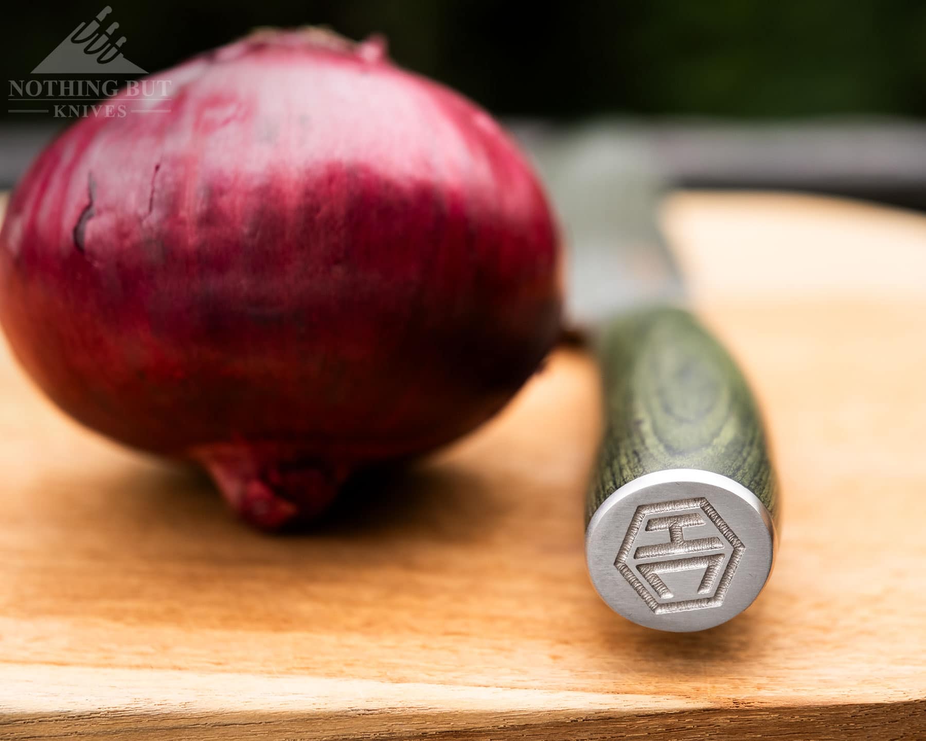 Hexclad Chef Knife Review - Is It Just Marketing Hype? - Image 4 A close-up of the HexClad chef knife handle pommel on a wood cutting board with a red onion in front of a blurry, green background.