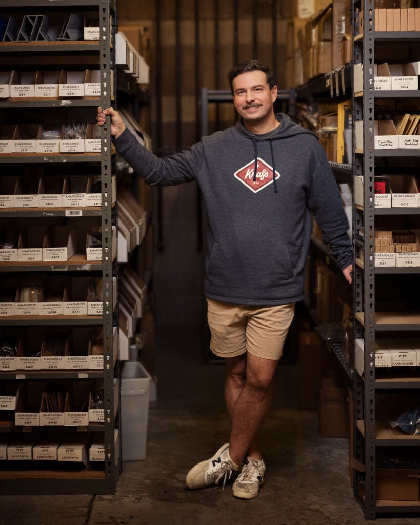 Blades N' Tales: Looking Inside The Knafs Rocket Ship - Image 4 A full length image of Austin Goetz of Knafs Knives standing between to supply shelves in the company warehouse.