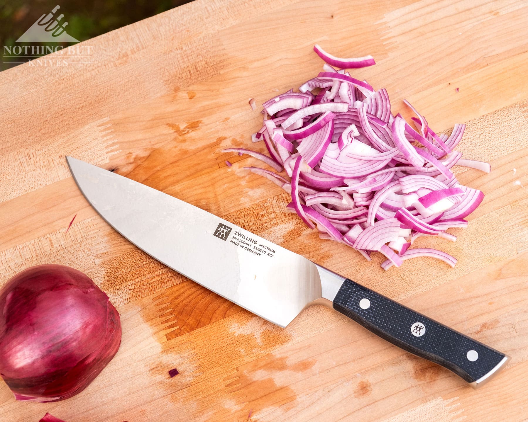 Zwilling Spectrum Chef'S Knife Review - Image 3 The Zwilling Spectrum laying on its side on a wooden cutting board with half of an onion and chopped onions.