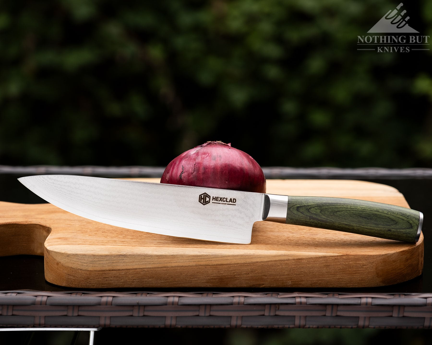 Hexclad Chef Knife Review - Is It Just Marketing Hype? - Image 1 A left side profile image of the HexClad Damascus chef knife next to a red onion on a wood cutting board in front of a blurry green background.