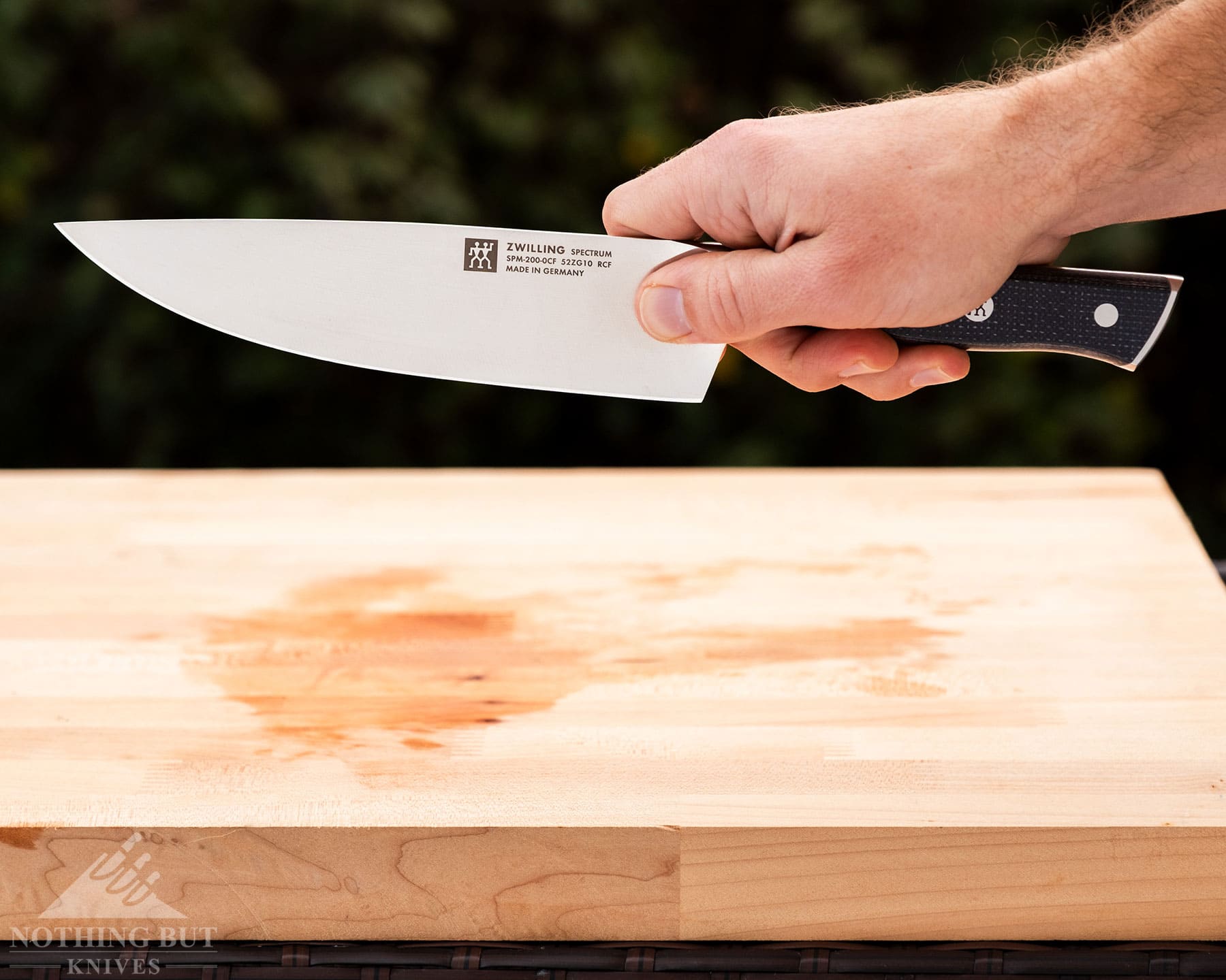 Zwilling Spectrum Chef'S Knife Review - Image 2 Zwilling Spectrum being held in the right hand above a wooden cutting board with greenery in the background.