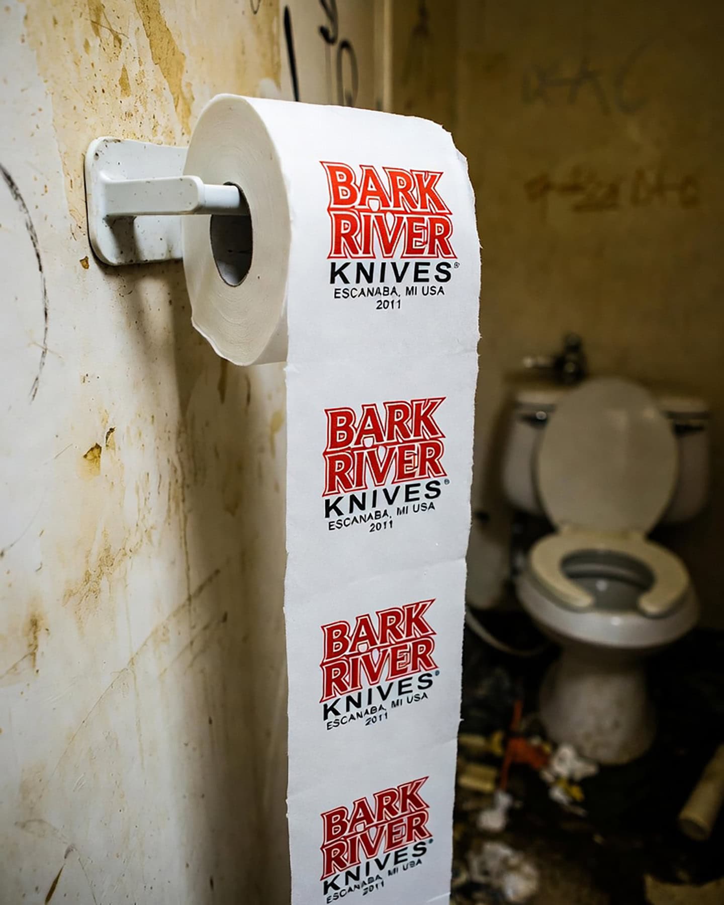 A roll of toilet paper with the Bark River Knives logo printed on it in a dirty public bathroom stall.

