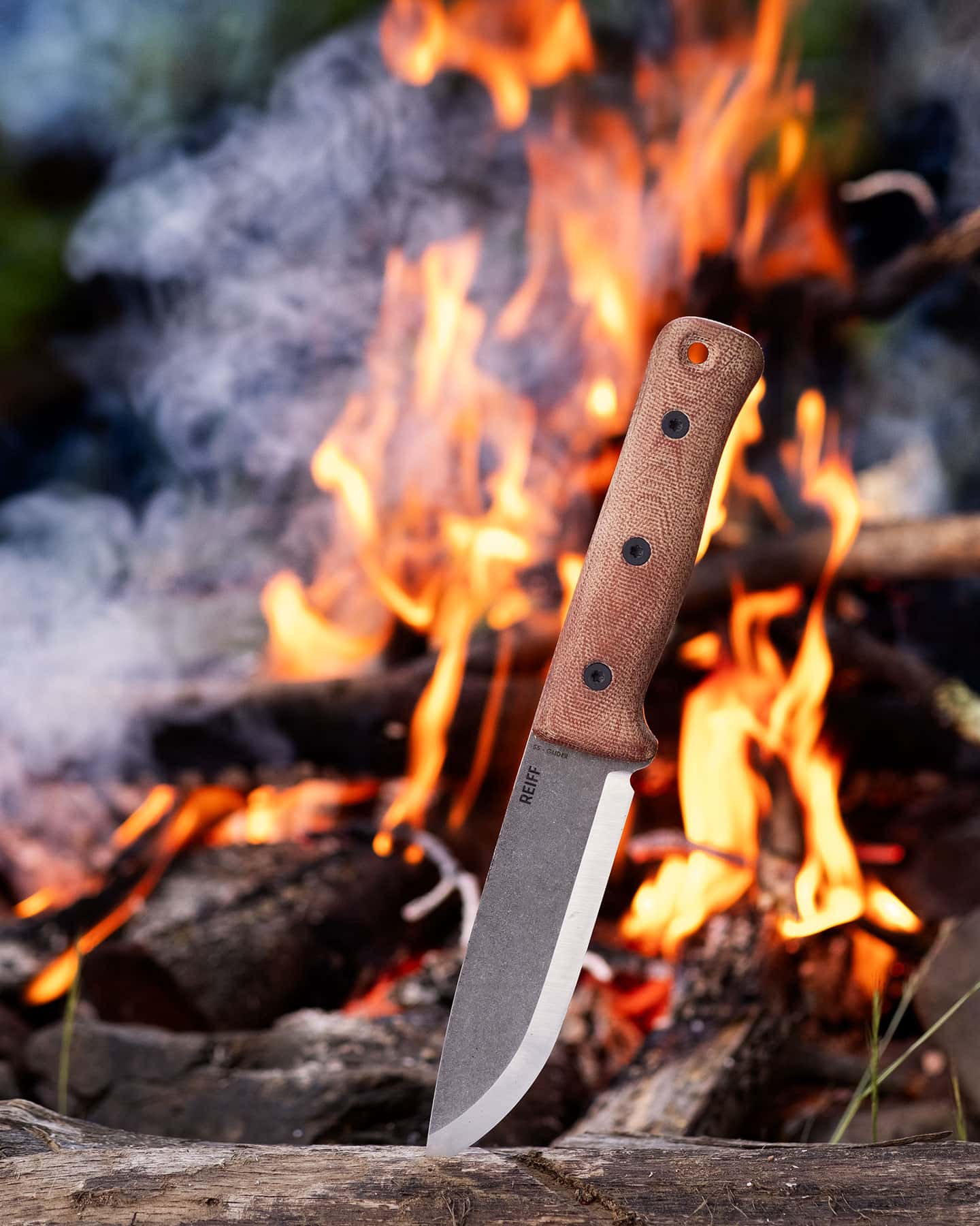 The Reiff F5 bushcraft knife sticking out of a log in front of a campfire.