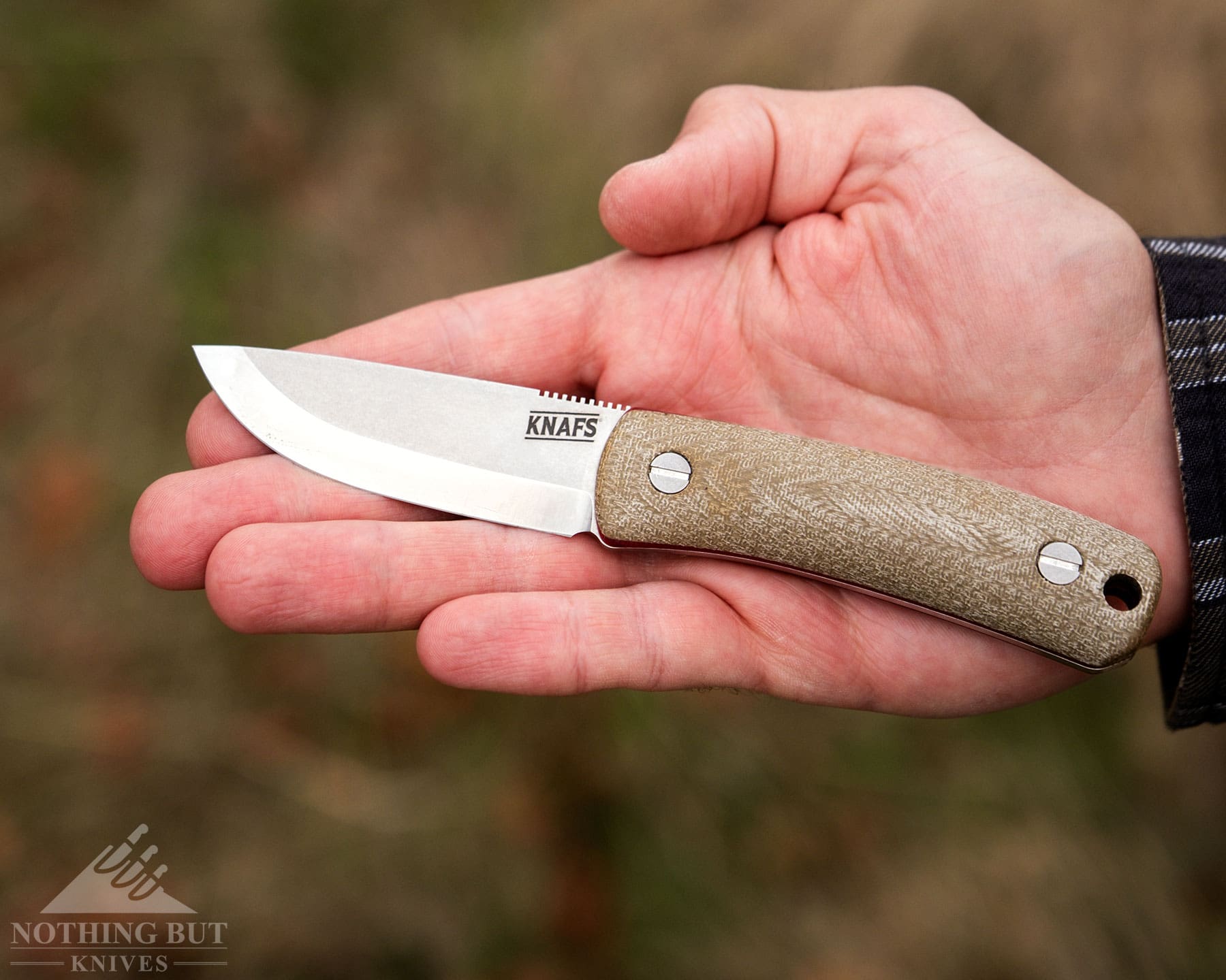 Knafs Lulu Review - Image 14 A fixed blade bushcraft knife being held in the right palm of a mans hand with forest in the background.