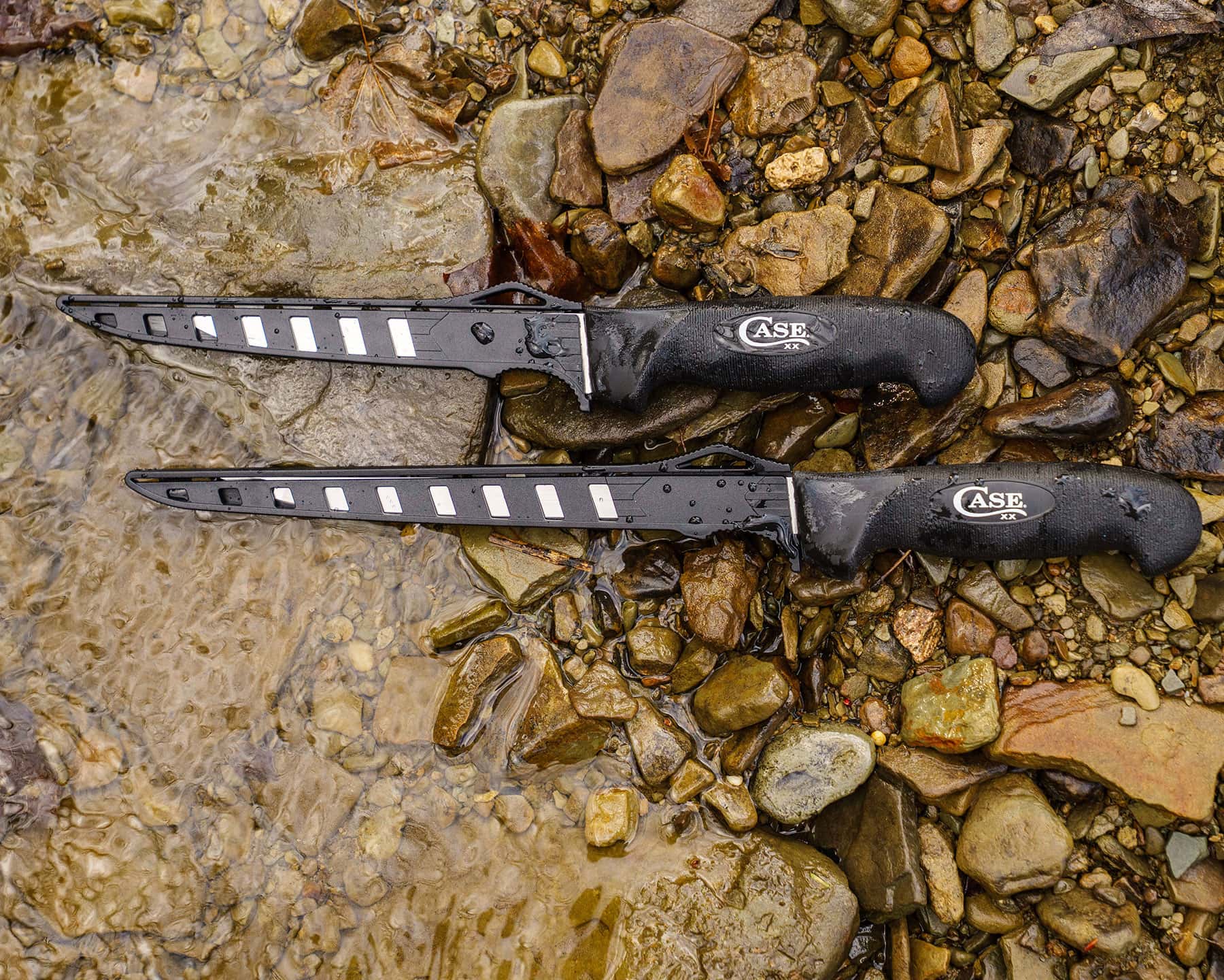 An overhead view of the six inch and eight inch Case fillet knives in their sheaths in the shallow waters of a freshwater beach. 