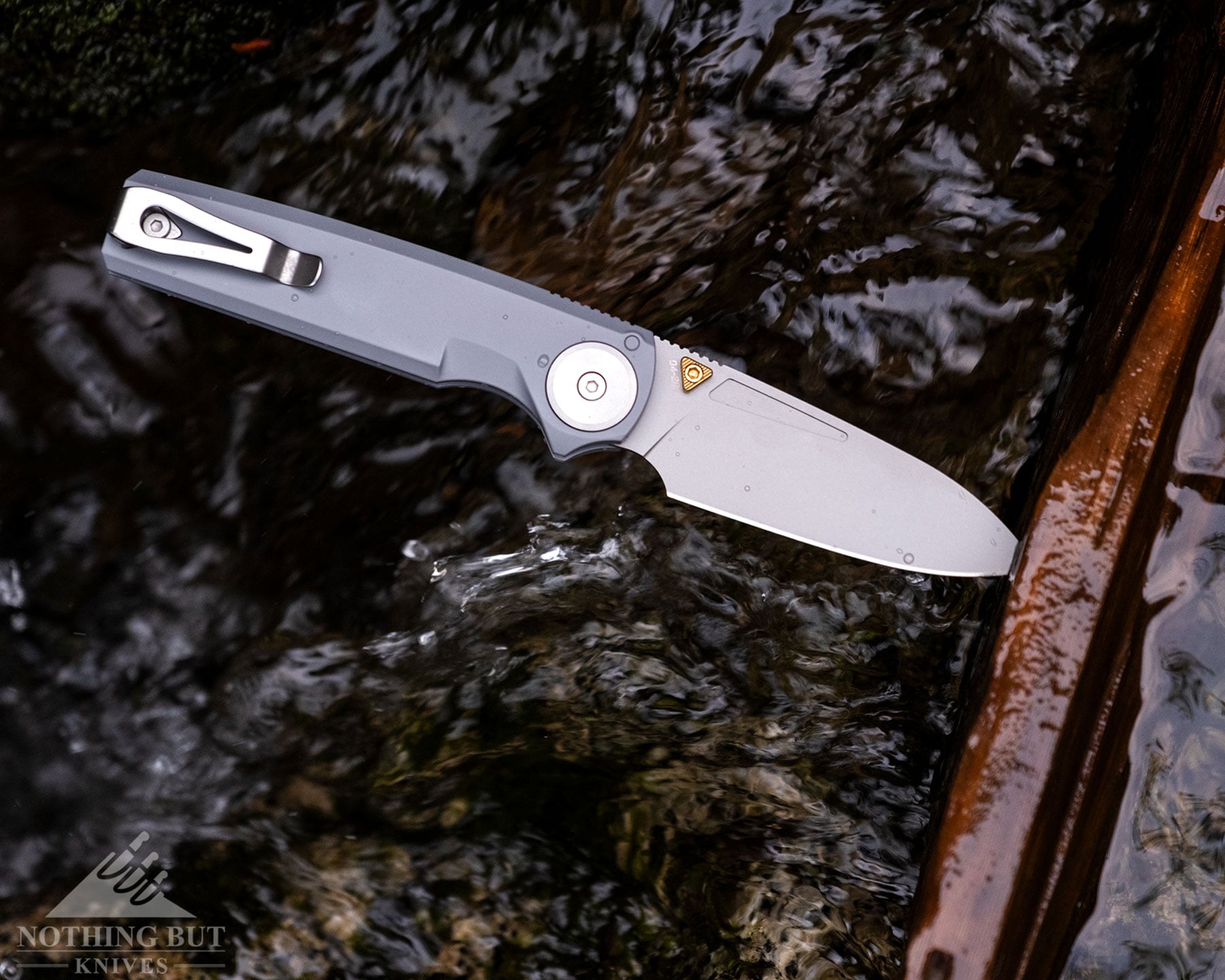Worksharp Rmx Thumbstud Knife Review - Image 2 An overhead view of the Work Sharp RMX folding knife with its blade ;locked in the open position sticking out of a log above a mountain creek.