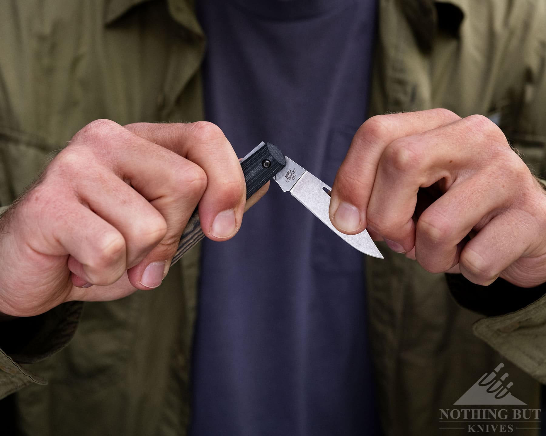 A close-up of a person in a black shirt and green jacket opening the W.R. Case Crossroads Barlow slip joint knife.