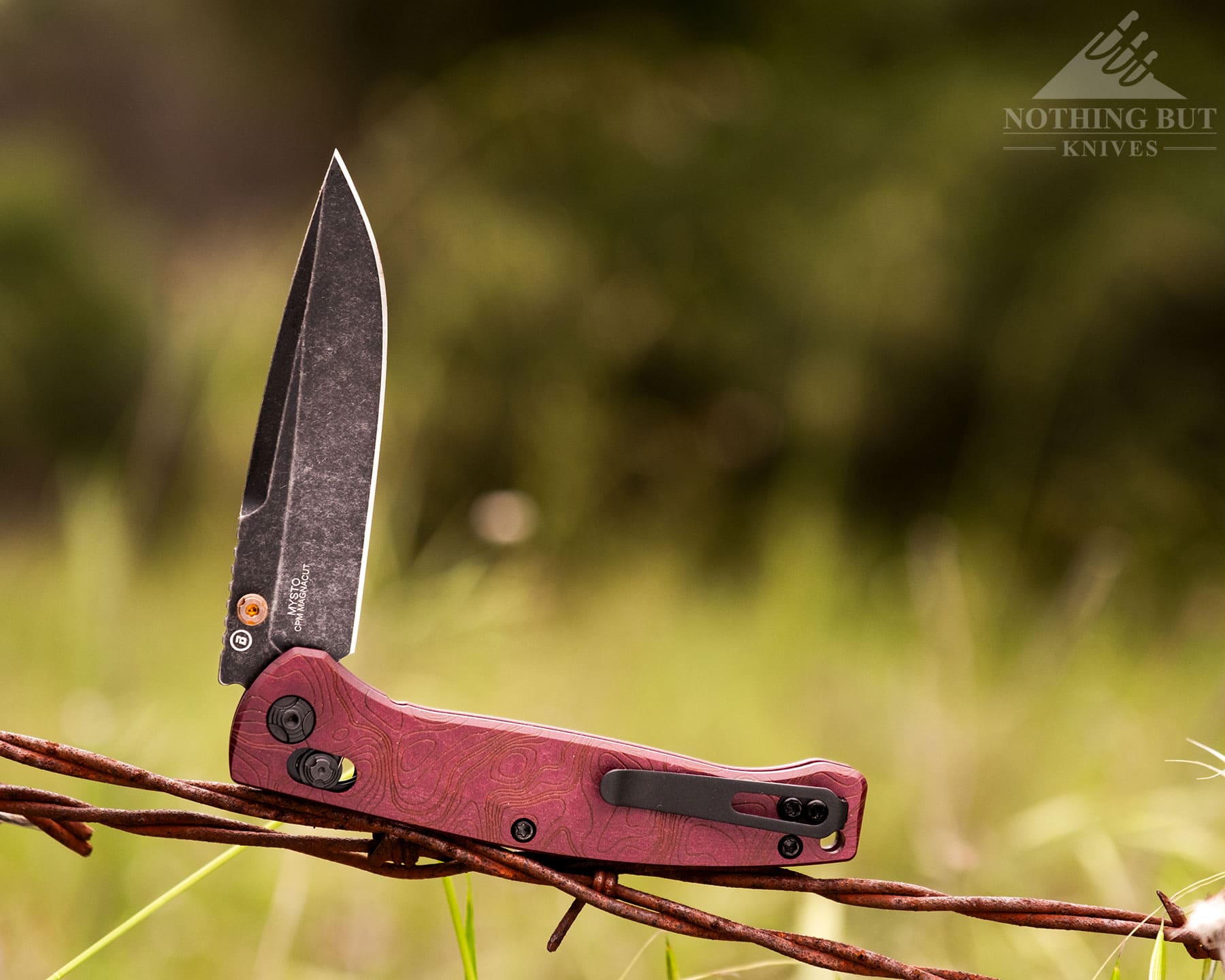 Hogue Mysto Review - Image 1 The Hogue Mysto folding knife with its blade in the half open position balanced on a strand of barbed wire in a green field.